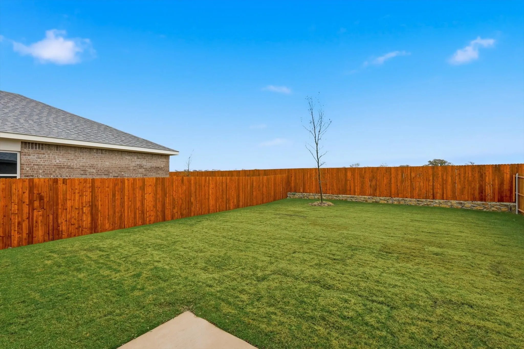 Expansive backyard featuring a new wood privacy fence, a manicured lawn, a concrete patio section, and a decorative stone retaining wall