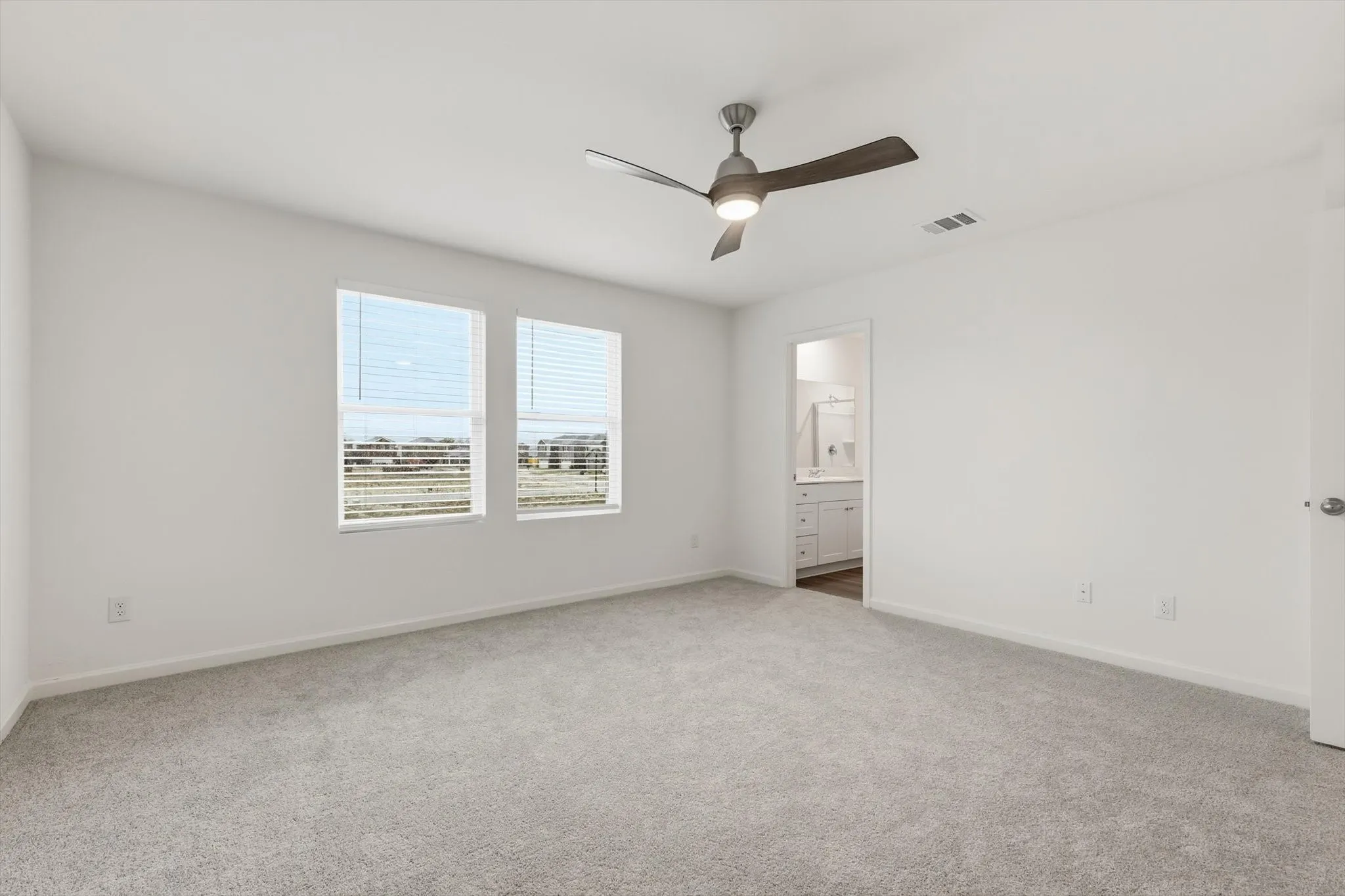 Spacious room with light gray carpeting, two large windows with horizontal blinds, and a contemporary ceiling fan with integrated lighting