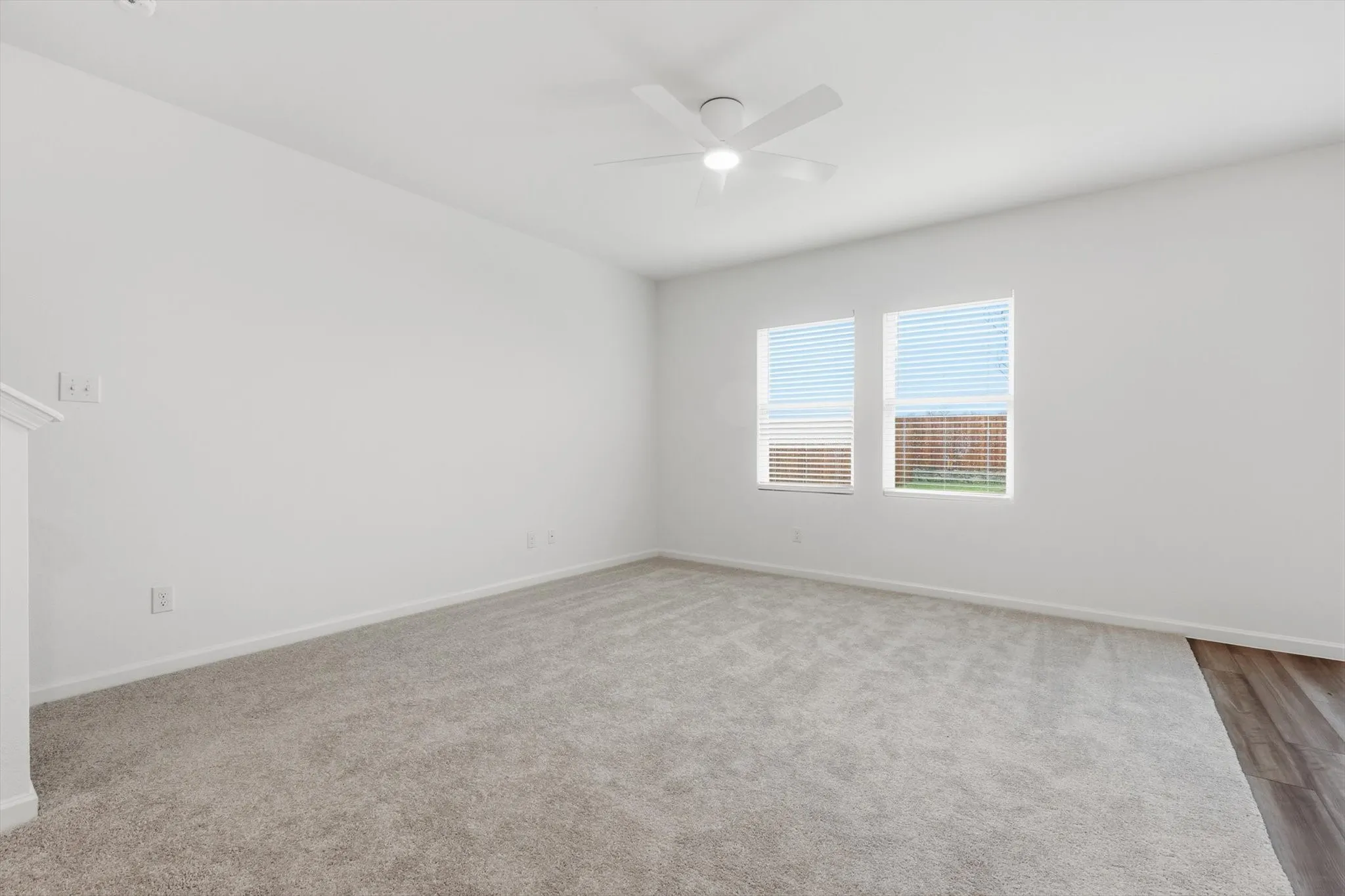Spacious room featuring light gray carpeting, crisp white walls, two windows with horizontal blinds, a white ceiling fan with integrated lighting, and a partial view of wood-finish flooring