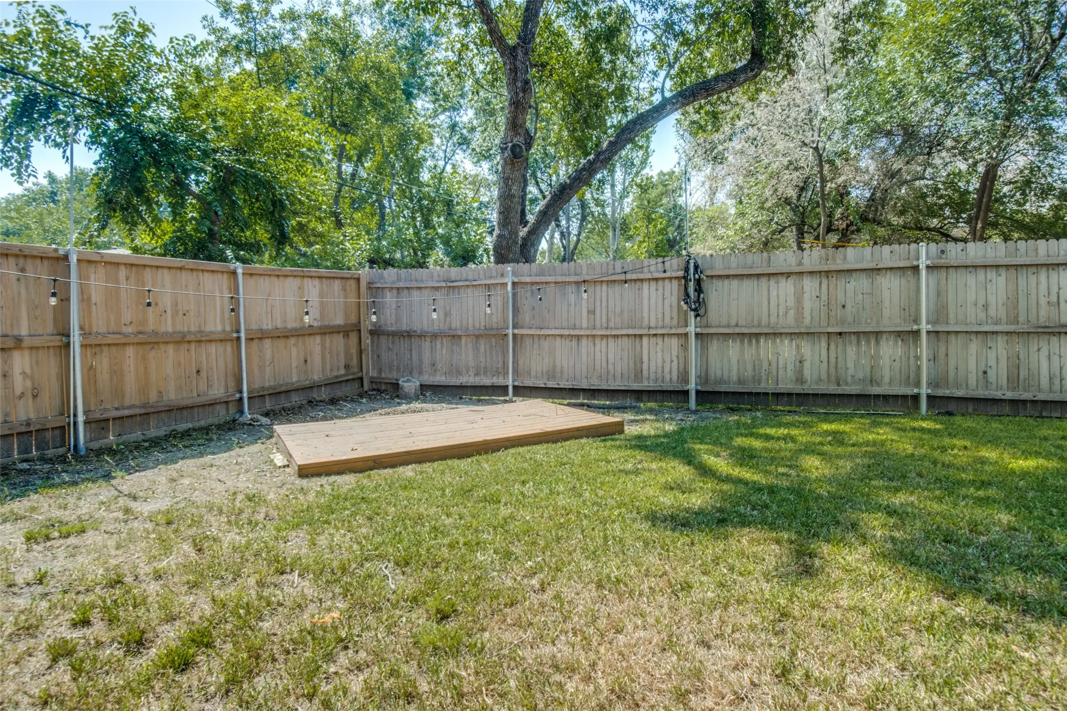 Fenced backyard featuring a wooden privacy fence, a grass lawn, a wood-plank platform, and string lighting