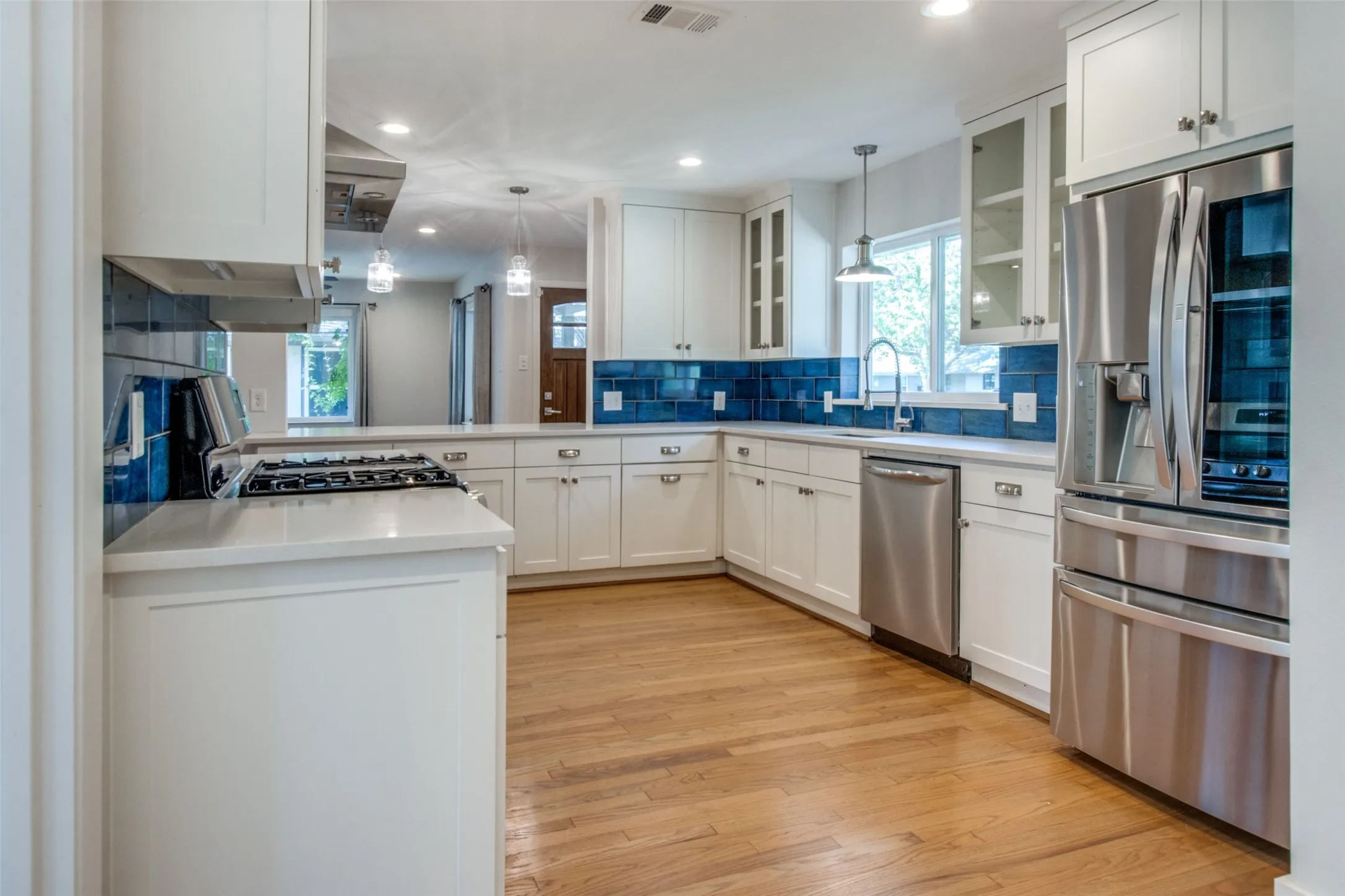 Spacious kitchen featuring white shaker-style cabinetry, a deep blue tile backsplash, stainless steel appliances, light-toned countertops, and wood-finish flooring