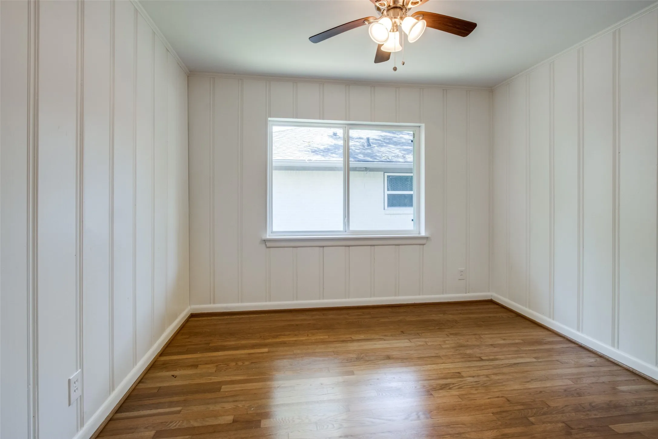 Room featuring wood-finish flooring, white board and batten walls, a single window, and a ceiling fan with integrated lighting