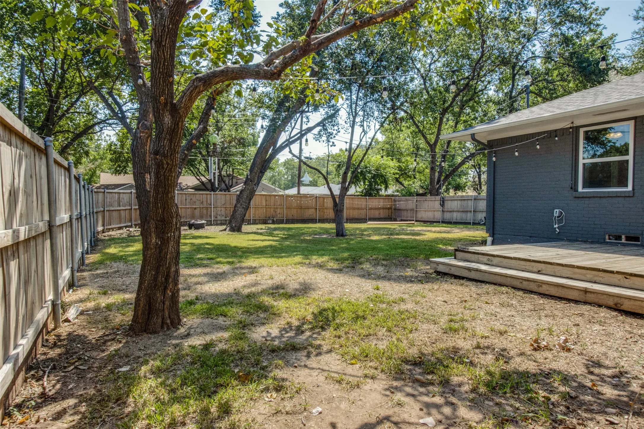 Expansive backyard featuring mature trees, a wooden privacy fence, a wooden deck with steps, and a dark-painted brick exterior with white trim