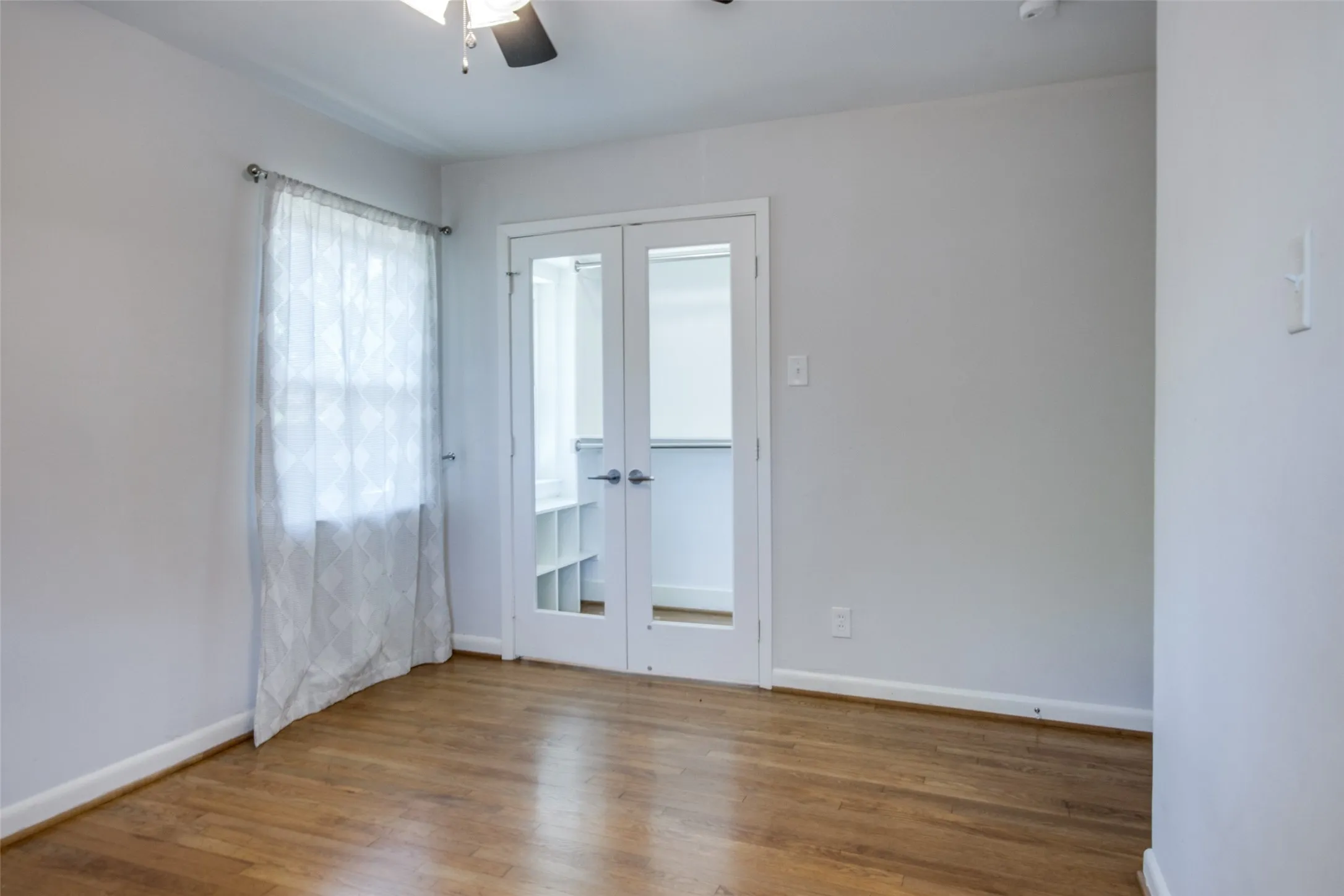 Wood-finish flooring throughout, featuring a ceiling fan, a window with sheer curtains, and double French doors opening to an additional organized space