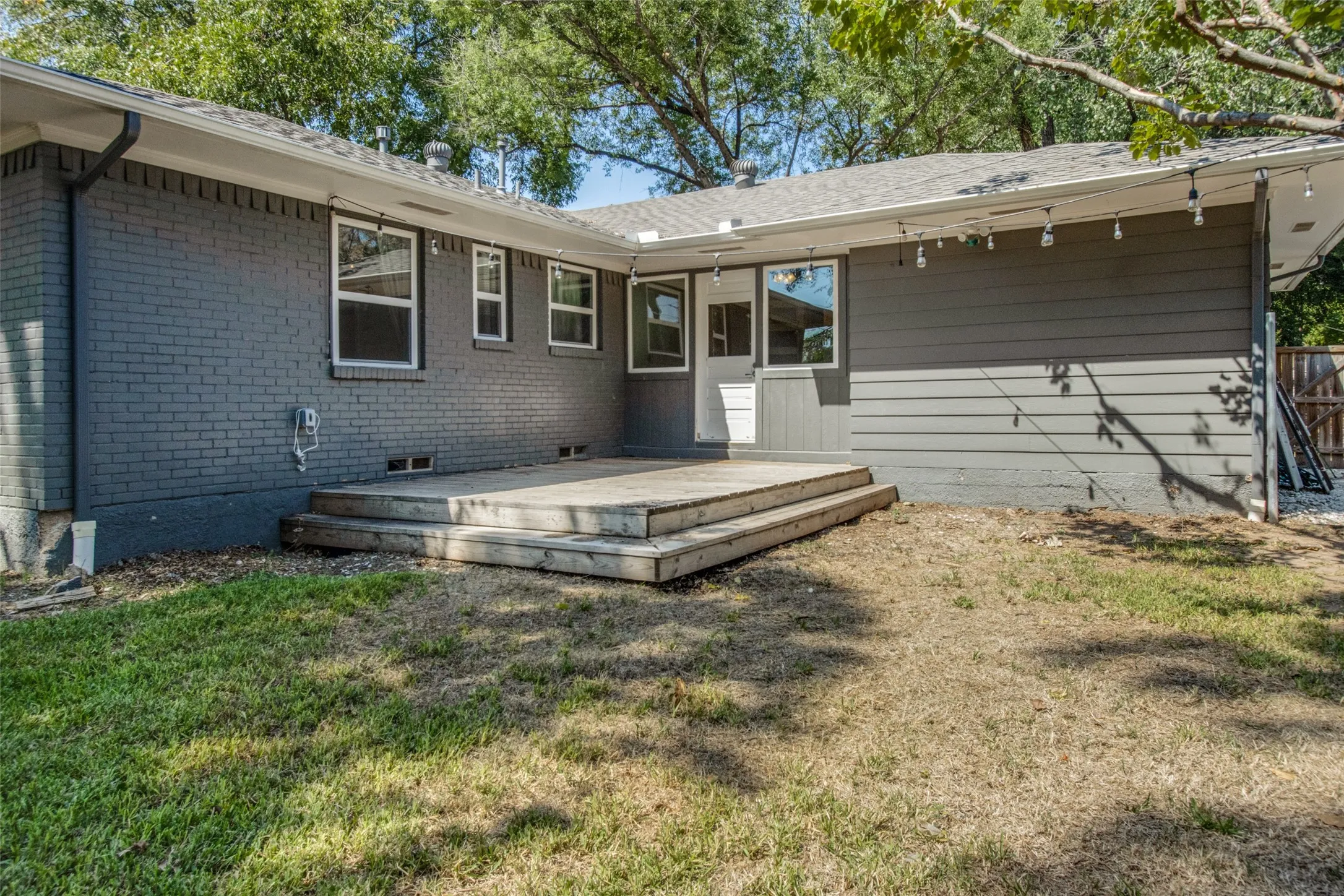 Rear exterior featuring a concrete patio with wood steps, grey brick siding, horizontal panel siding, multiple windows, and a white exterior door