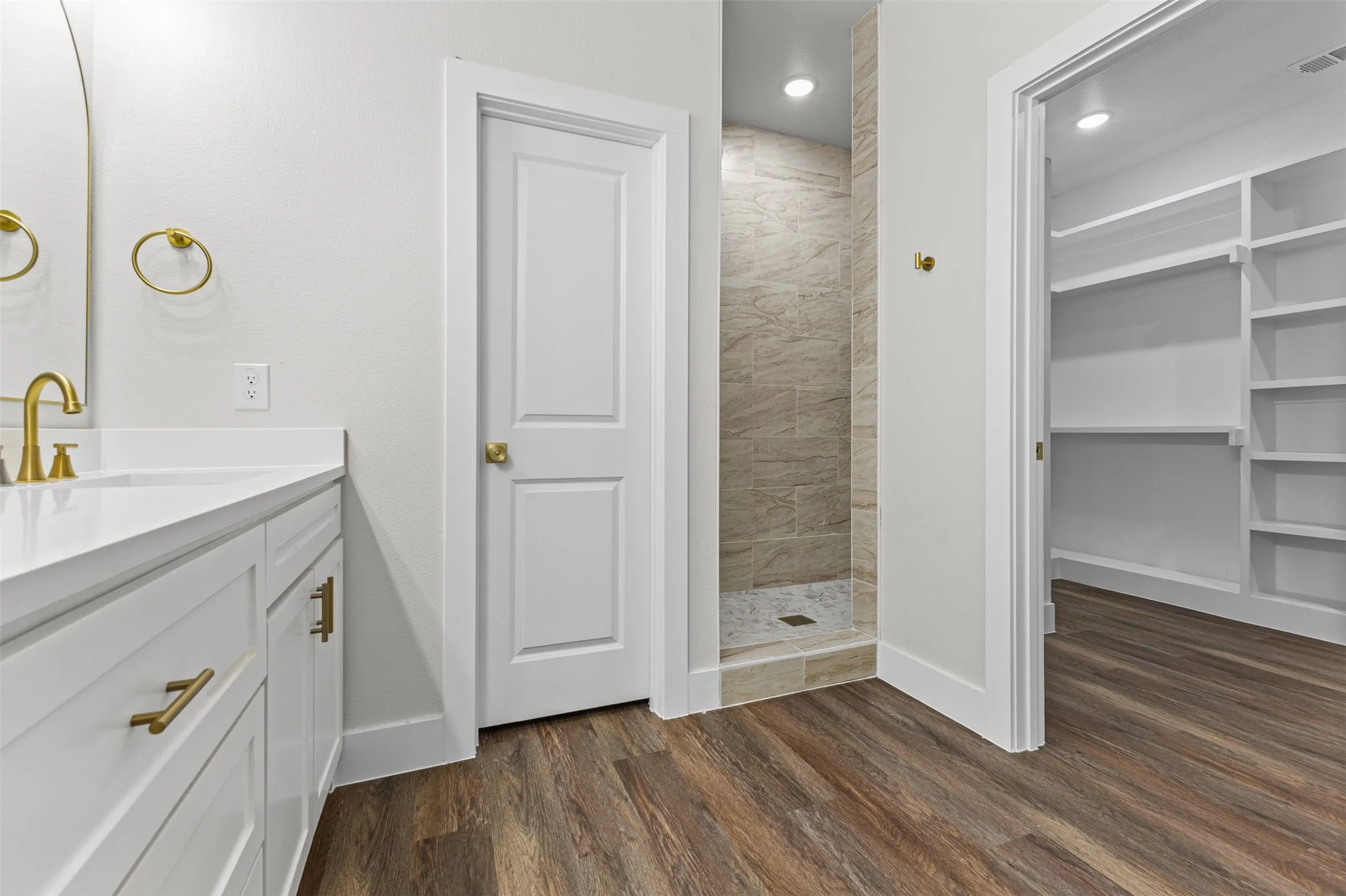 Bathroom featuring a white vanity with gold-tone hardware, a tiled walk-in shower, wood-finish flooring, and a separate closet with built-in shelving