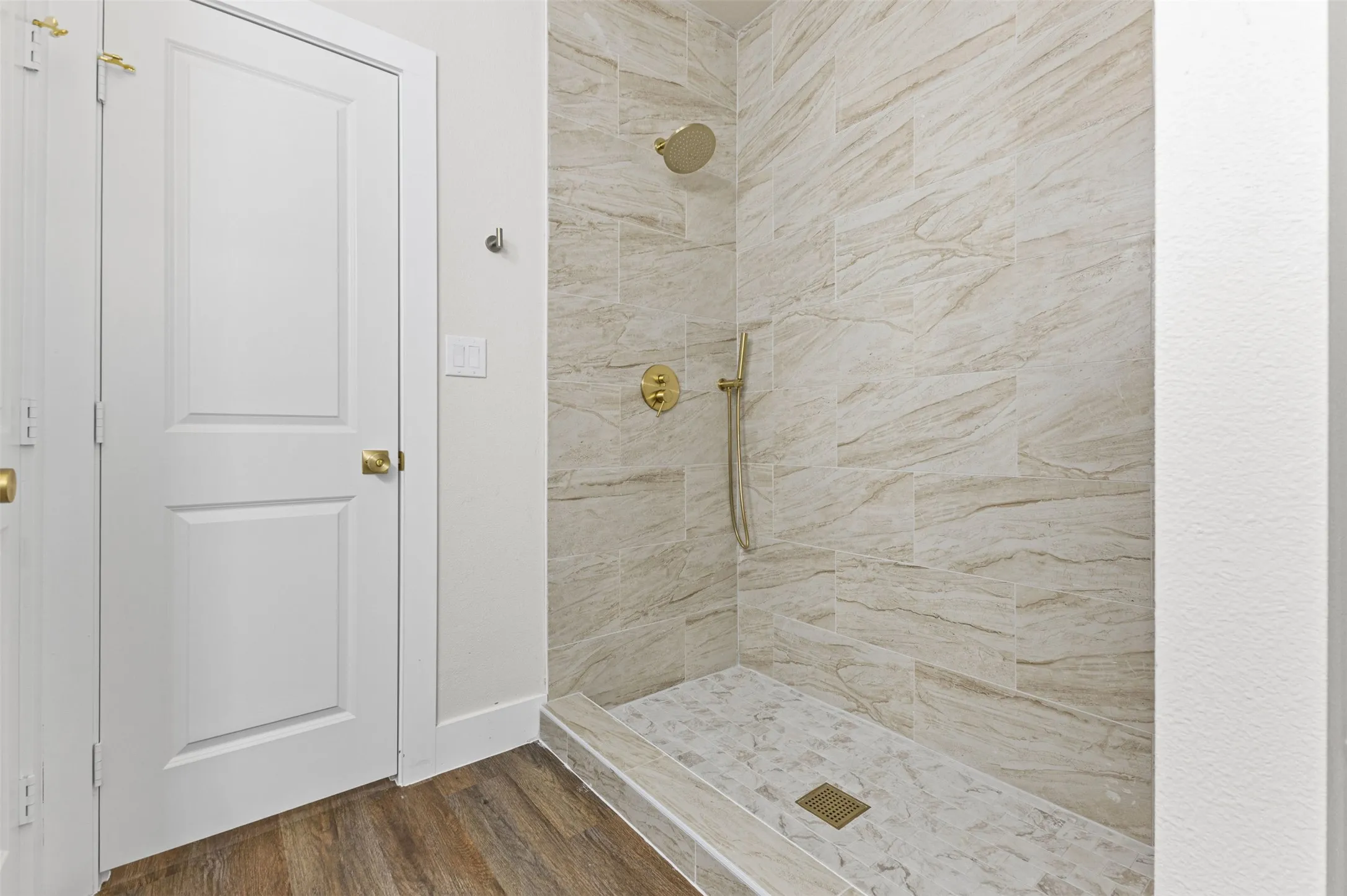 Walk-in shower featuring gold-finish fixtures, large format wall tiling, a tiled floor with a linear drain, and a contrasting wood-finish flooring outside the shower