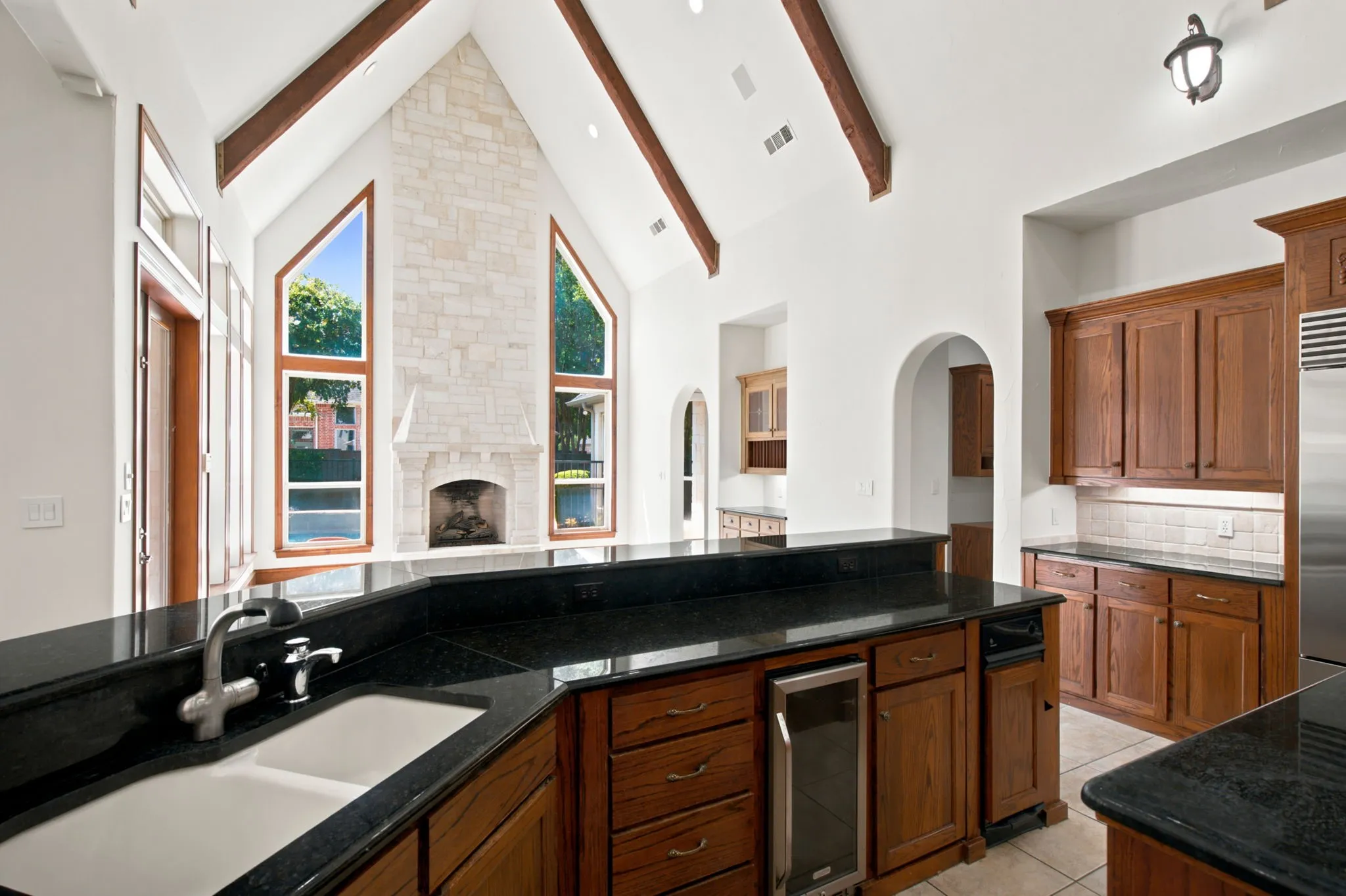 Vaulted ceiling with exposed wood beams, stone fireplace, custom wood cabinetry, dark stone countertops, and built-in wine cooler