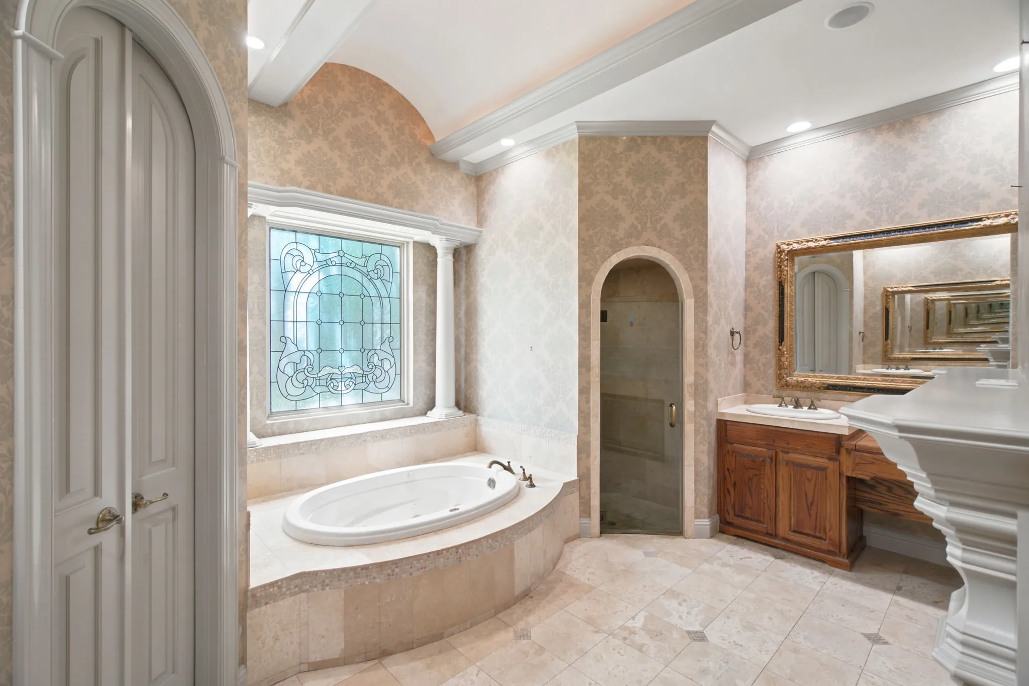 Recessed soaking tub with surrounding stone tile, custom stained-glass window, arched entry, standalone shower with glass enclosure, and wood-finish vanity