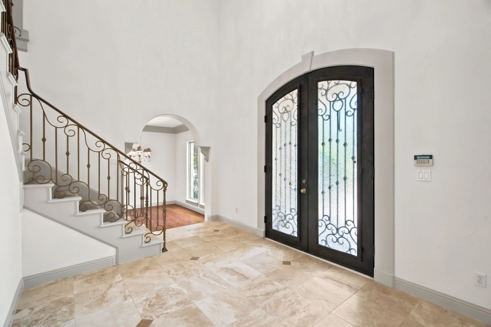 Arched double entry doors with decorative ironwork, a grand staircase with ornate wrought iron balusters, light-toned tile flooring with contrasting accents, and an arched doorway leading to a room with wood-finish flooring