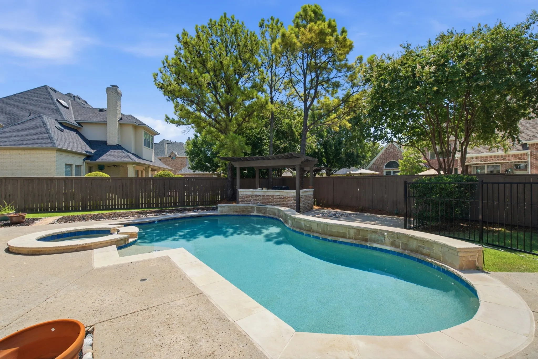 Backyard featuring a freeform pool, integrated spa, surrounding concrete decking, and a stone-accented outdoor cooking area with pergola