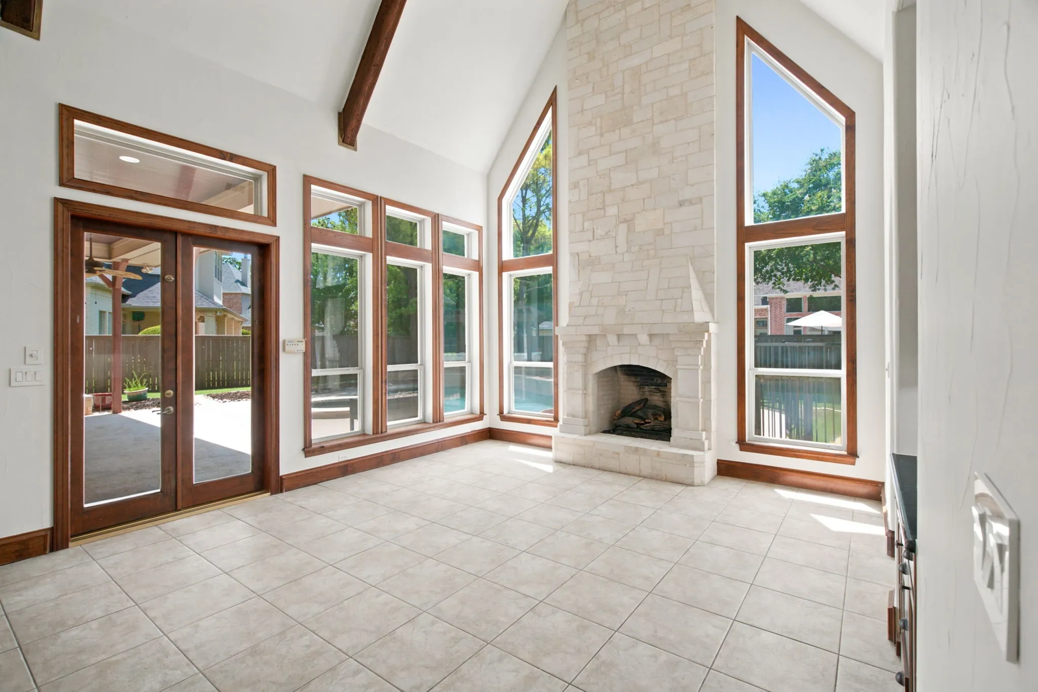 Vaulted ceiling featuring exposed beams, floor-to-ceiling stone fireplace, expansive windows with wood trim, and tile flooring