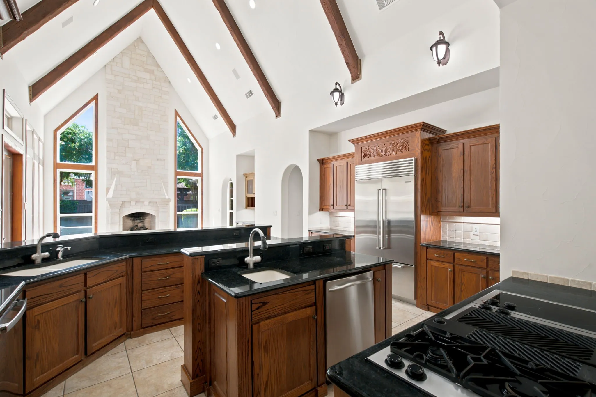 Vaulted ceilings with exposed wood beams, stone fireplace, custom wood cabinetry, integrated stainless steel appliances, and dark countertops