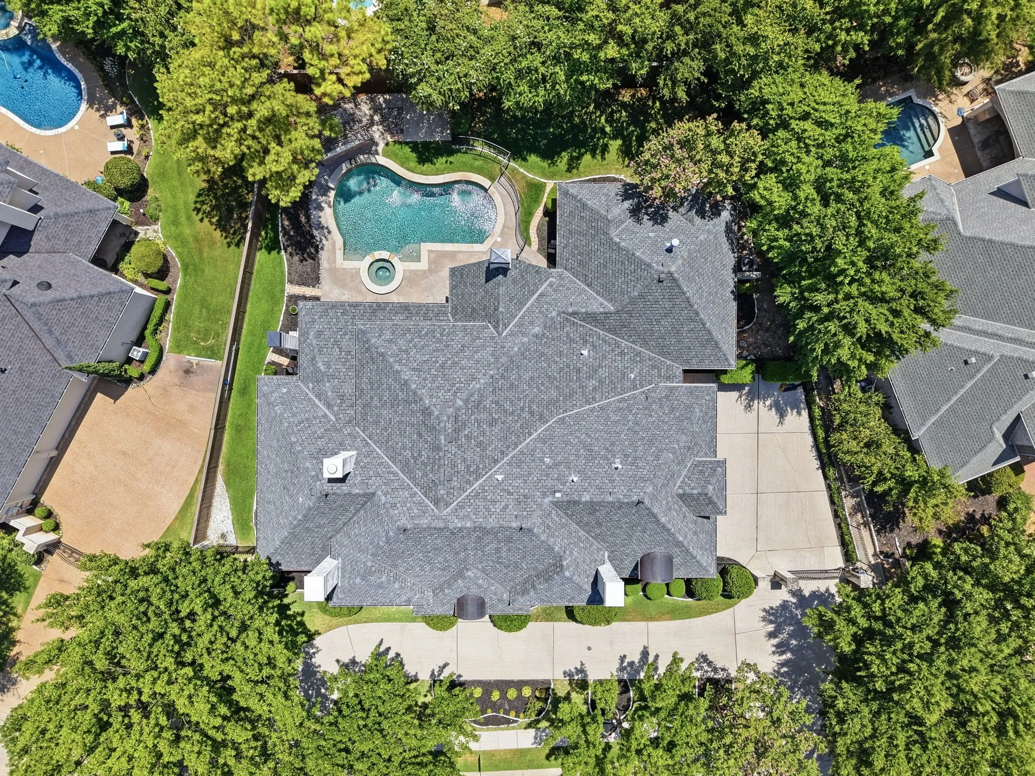 Expansive property featuring a multi-gabled roofline, concrete driveway, lush landscaping, and a curvilinear swimming pool with an integrated spa