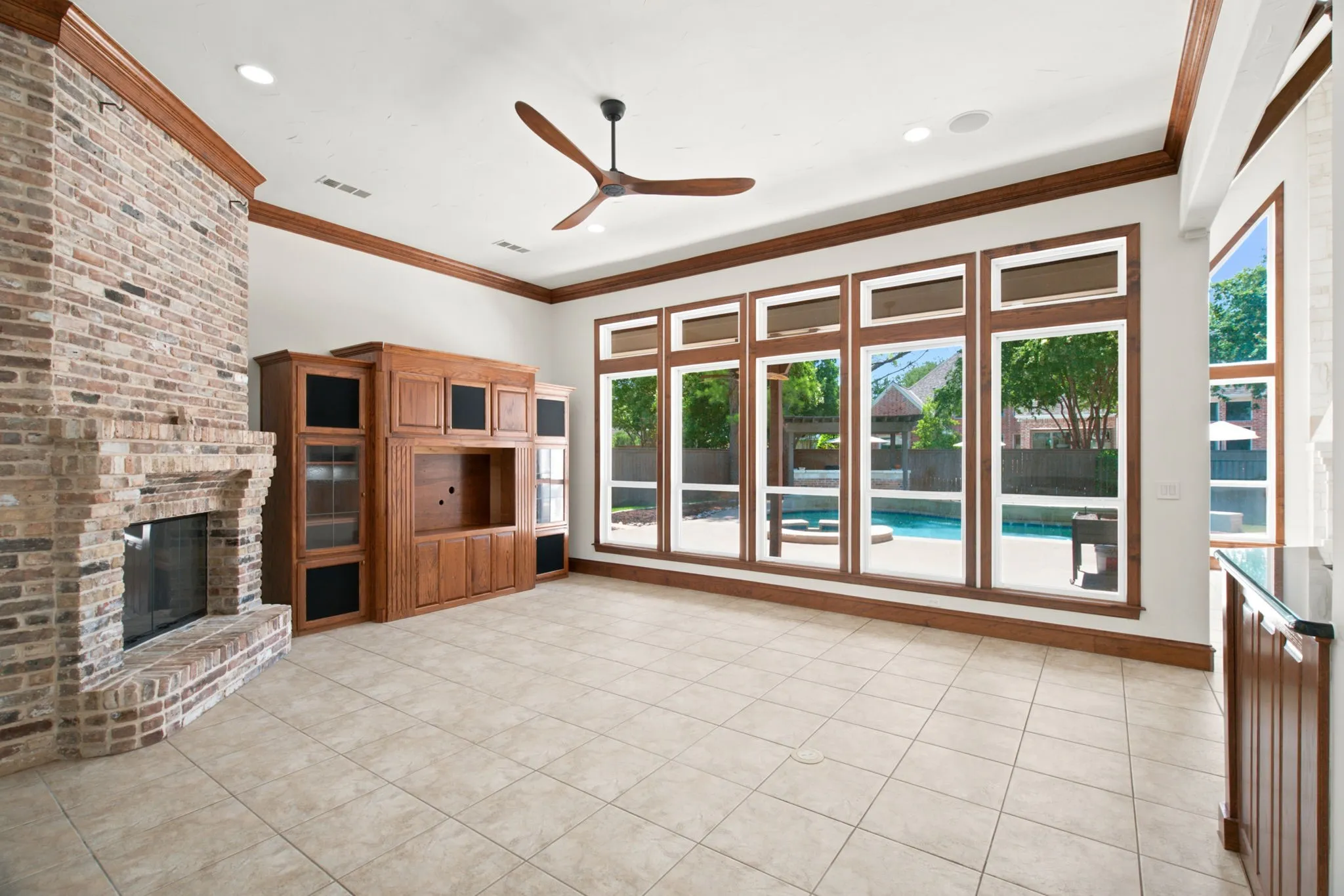 Spacious room featuring a brick fireplace, built-in entertainment center, tile flooring, a ceiling fan, and large windows with exterior pool access