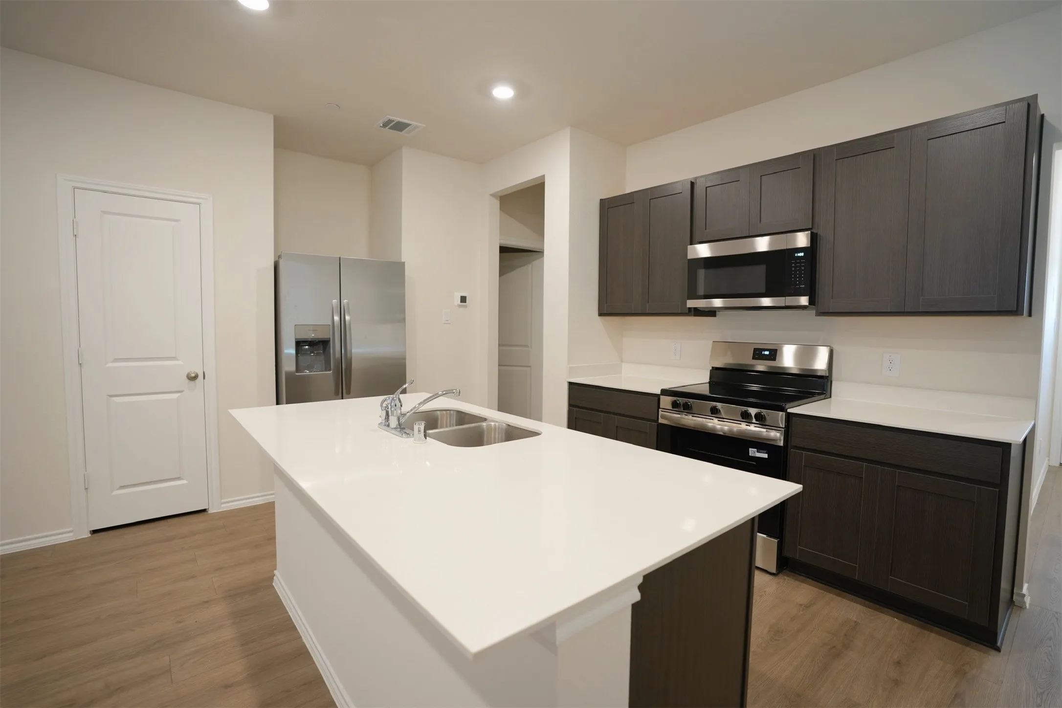 Modern kitchen featuring a central island with a double basin sink, dark wood-finish cabinetry, stainless steel appliances, light countertops, and wood-finish flooring