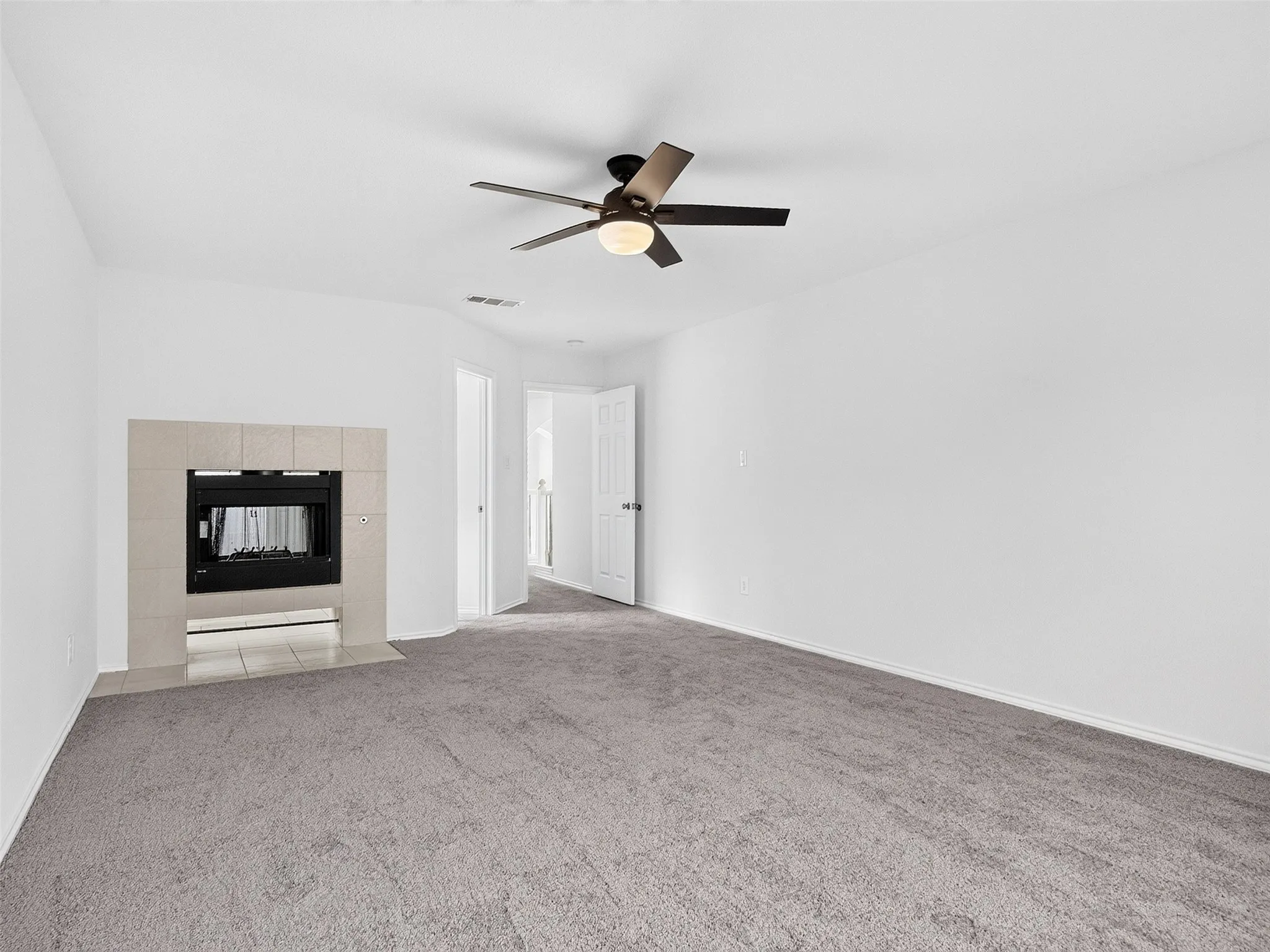 Carpeted room featuring a ceiling fan, a tiled fireplace with an integrated firebox, white walls, and a white door