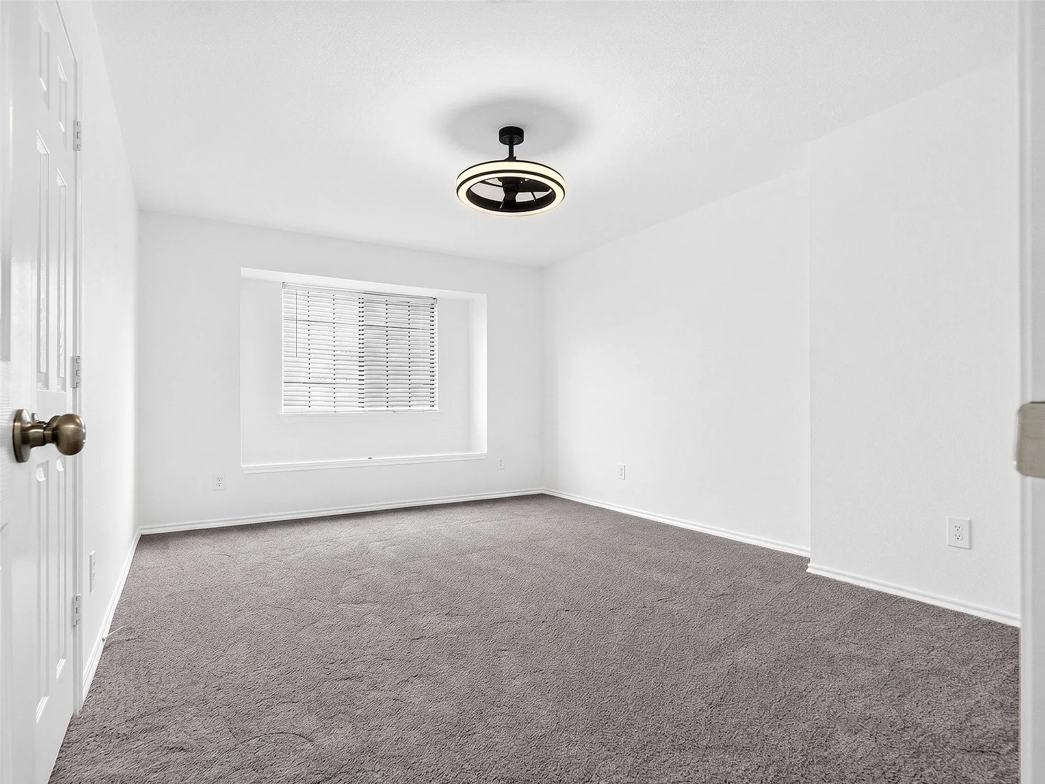 Neutral-toned carpeting extends throughout this room featuring a window with integrated blinds, a modern ceiling light fixture, white trim, and a paneled door with a brass-finish doorknob