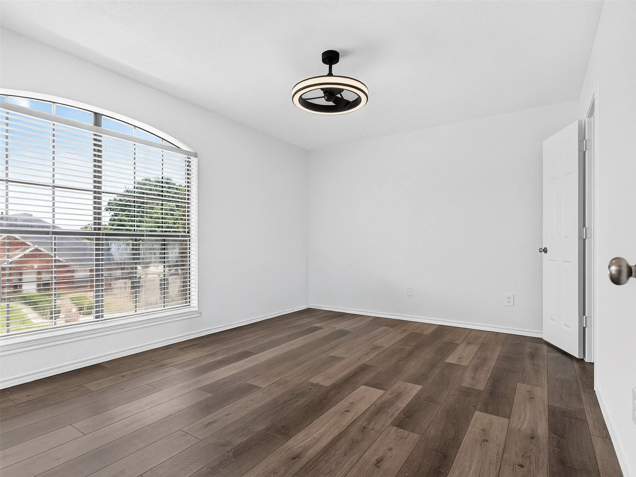 Bright interior space featuring a large arched window with horizontal blinds, wood-finish flooring, a contemporary ceiling fan with integrated lighting, and a white paneled door