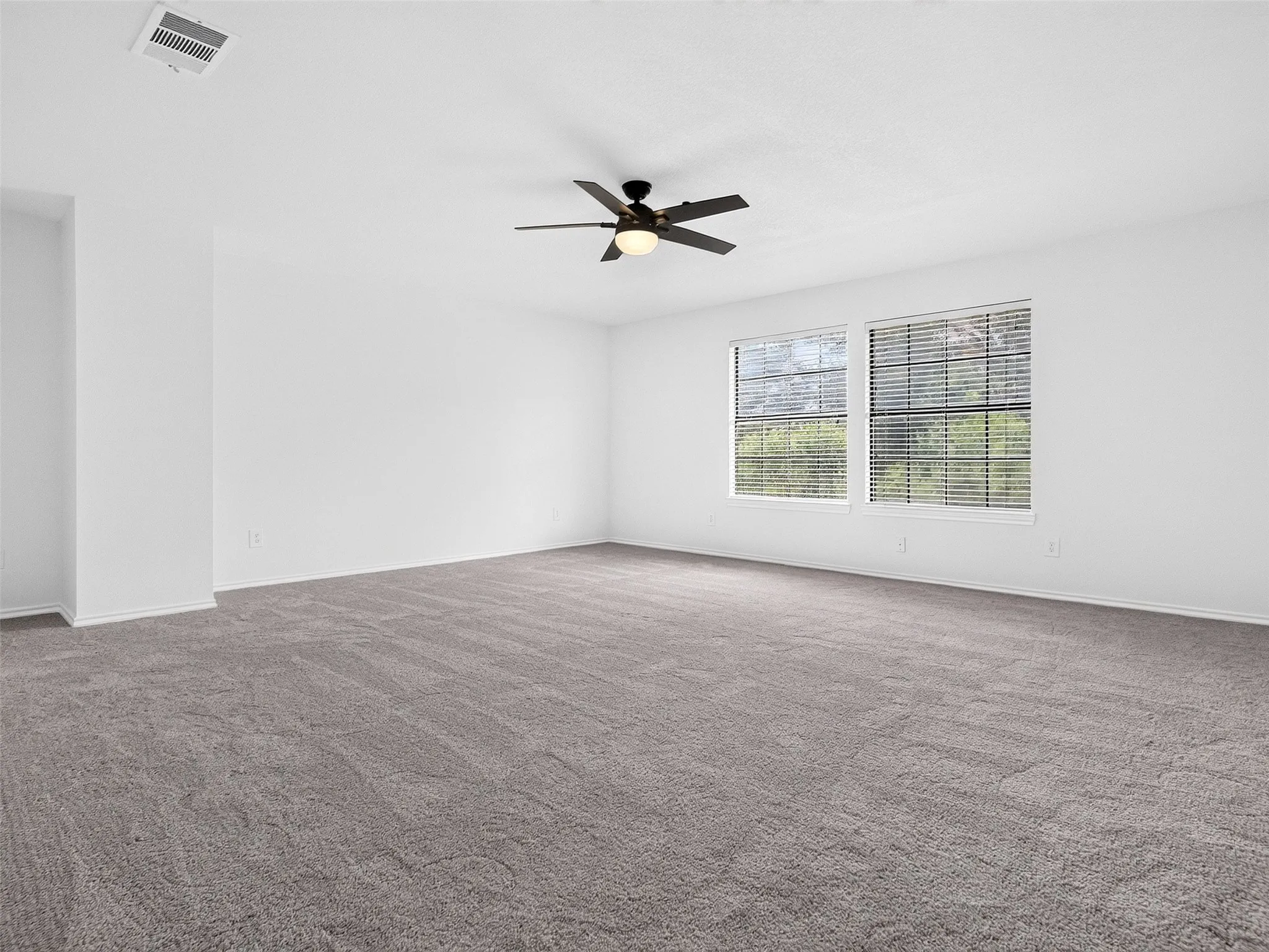 Spacious room featuring neutral carpeting, white walls, two large windows with blinds, a ceiling fan with integrated lighting, and a visible ventilation register