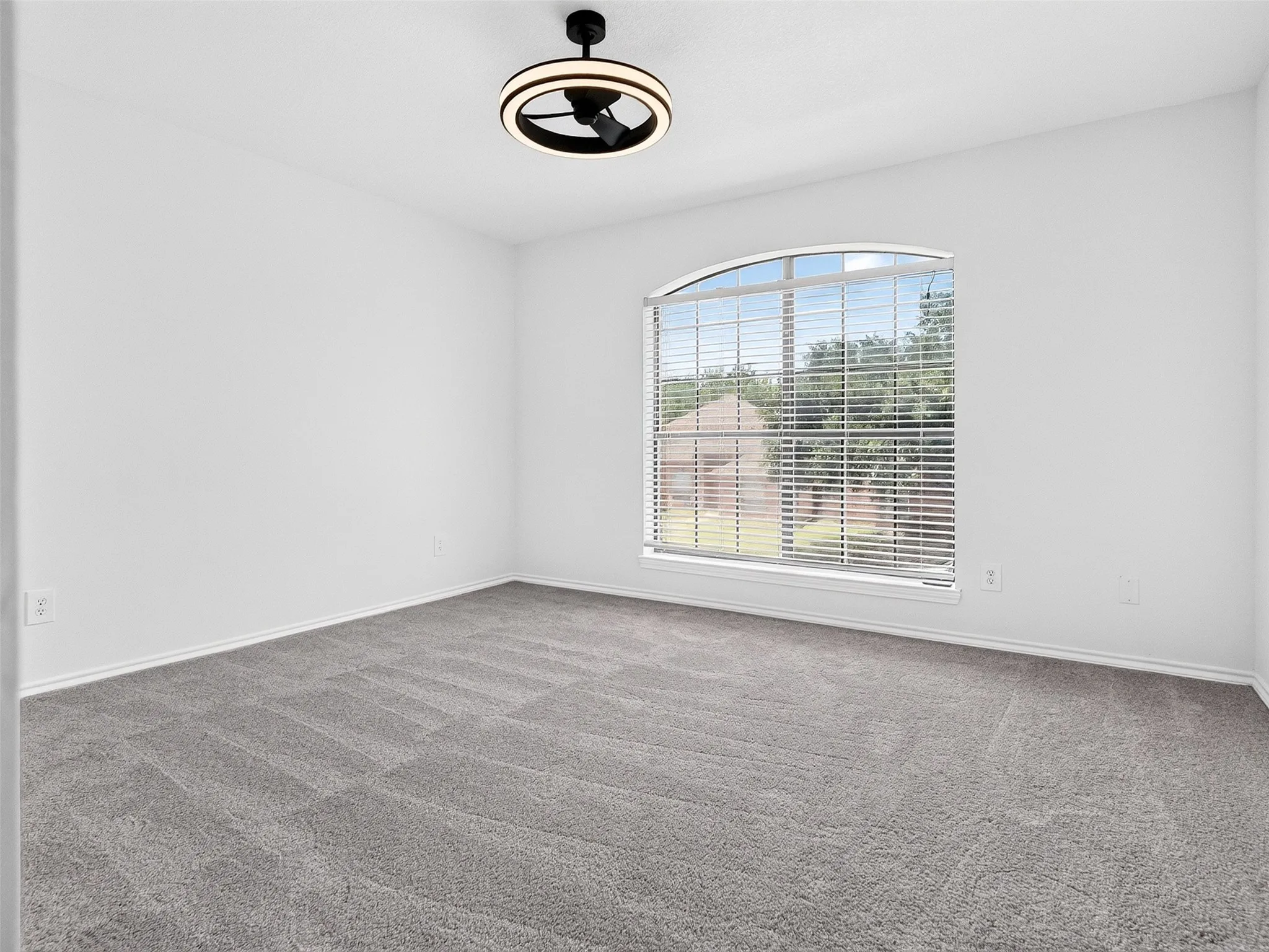 Spacious room featuring gray carpeting, an arched window with horizontal blinds, and a contemporary ceiling fan with integrated lighting