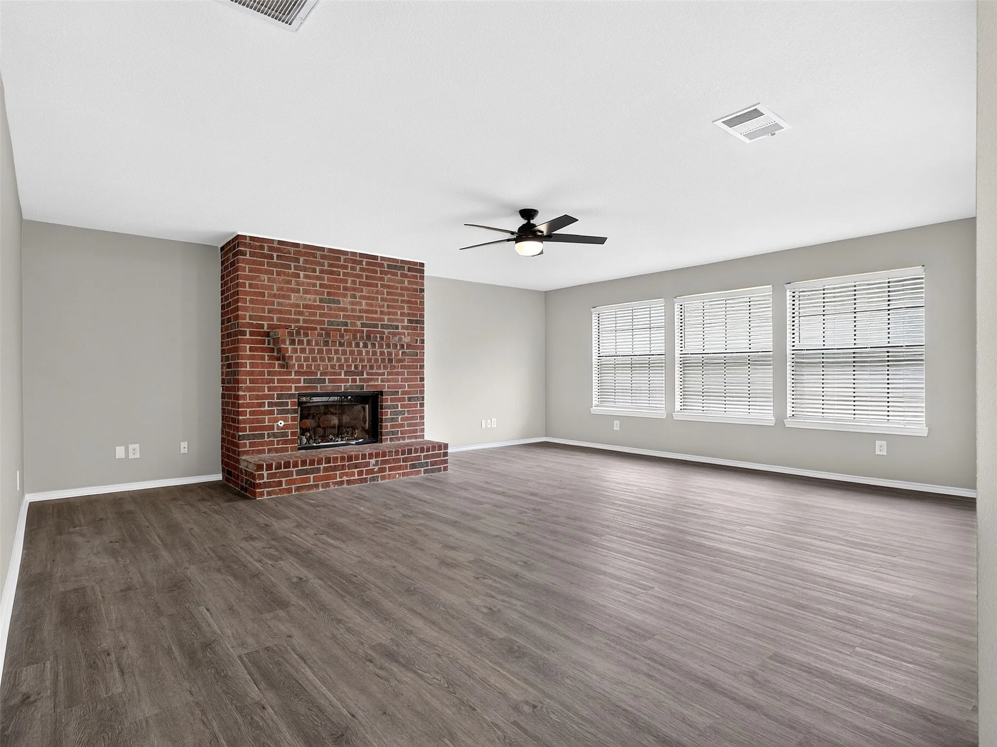 Spacious room featuring a prominent brick fireplace with a raised hearth, wood-finish flooring, light gray walls, and multiple windows with blinds