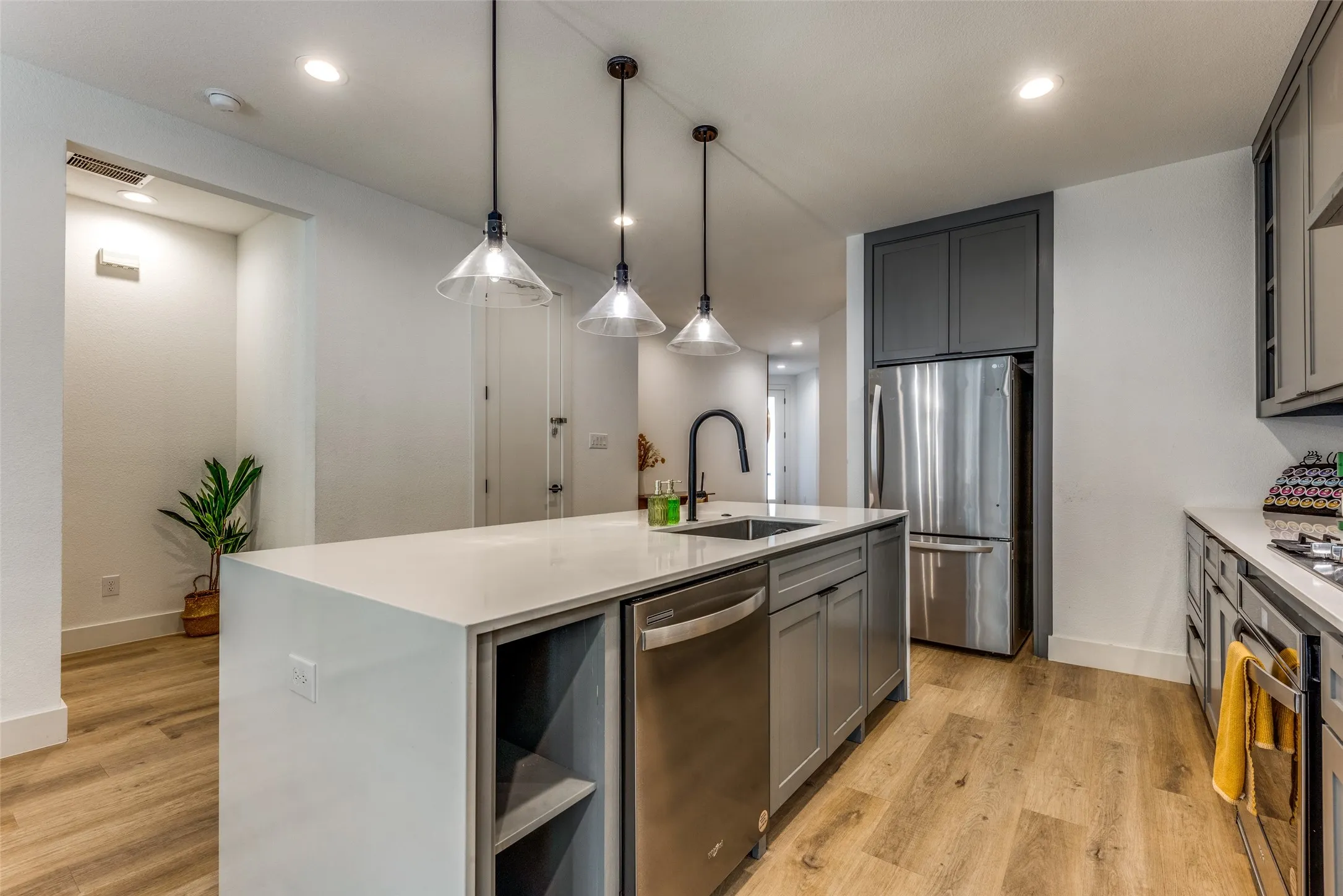 Modern kitchen featuring an island with a light-toned countertop, stainless steel appliances, dark gray cabinetry, three pendant lights, and wood-finish flooring