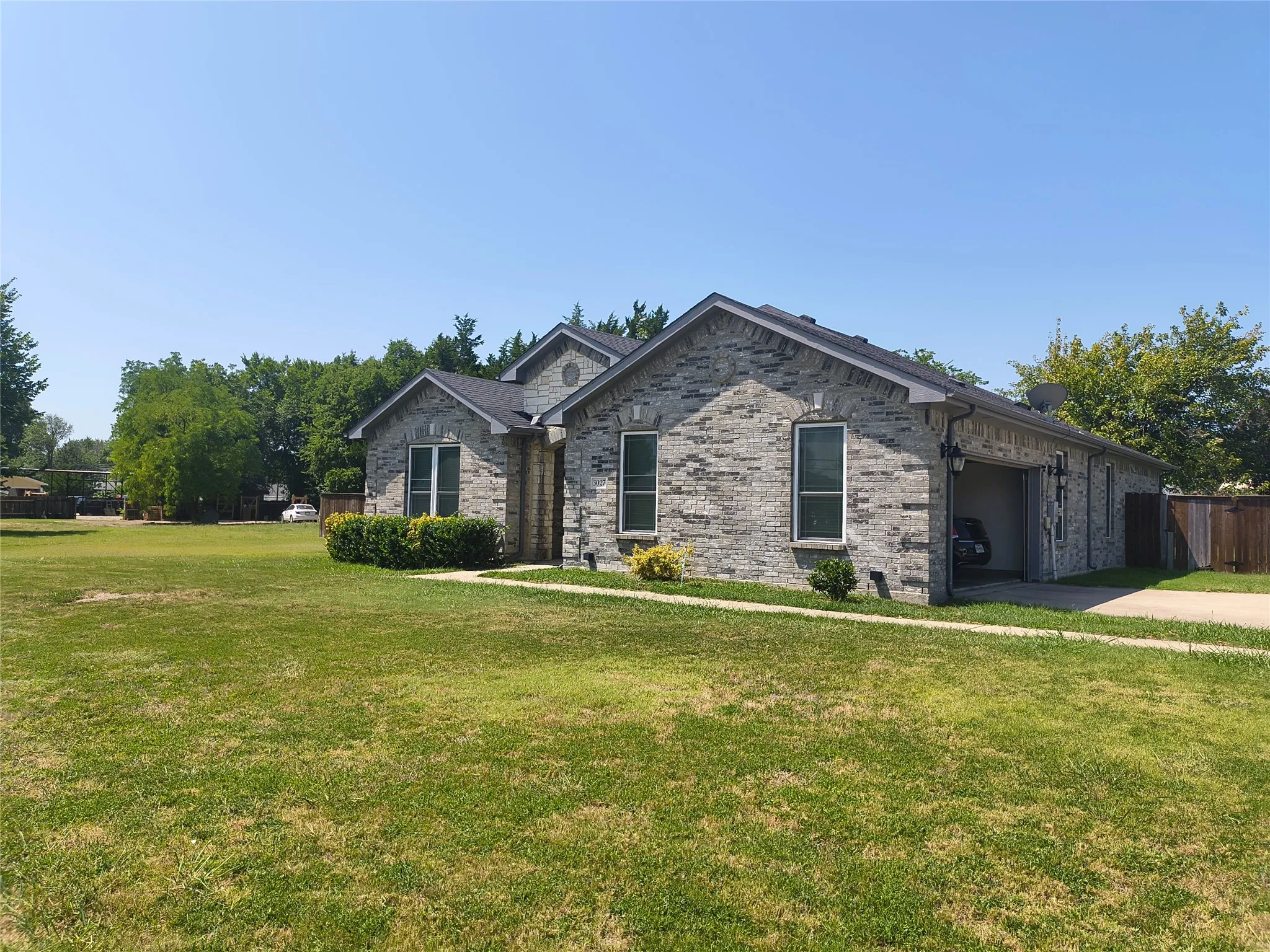 Grey brick exterior with multiple window openings, an attached garage, a dark asphalt shingle roof, a concrete driveway, and front landscaping