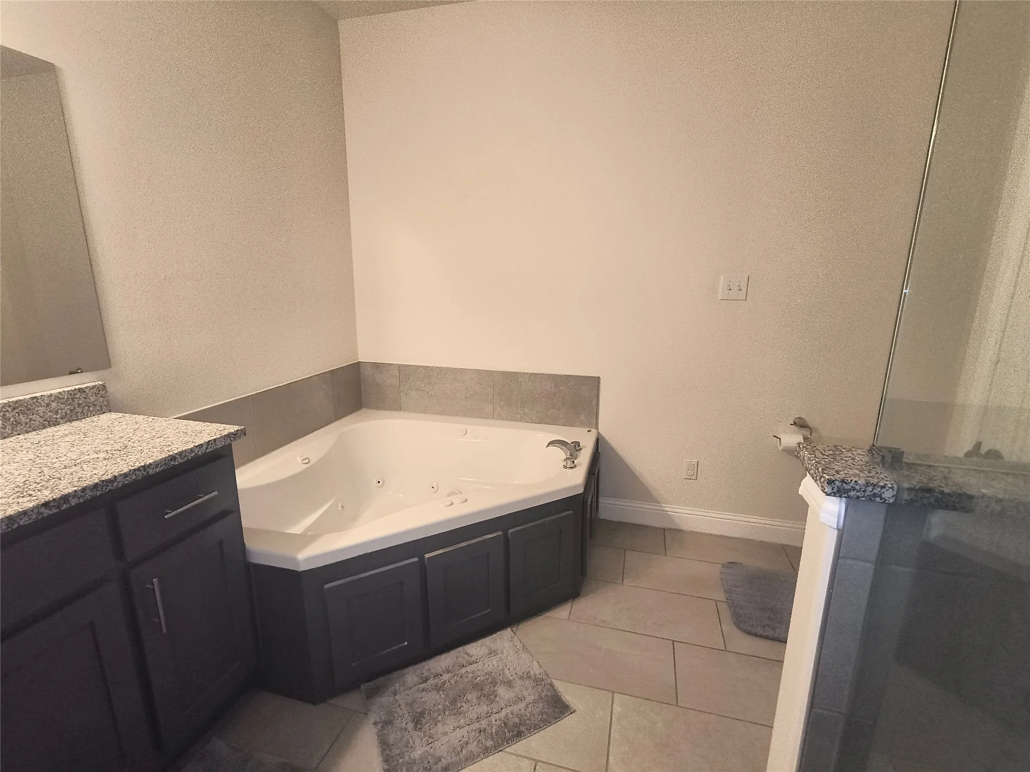 Corner jetted tub with dark cabinetry surround, tile flooring, and a granite-topped vanity