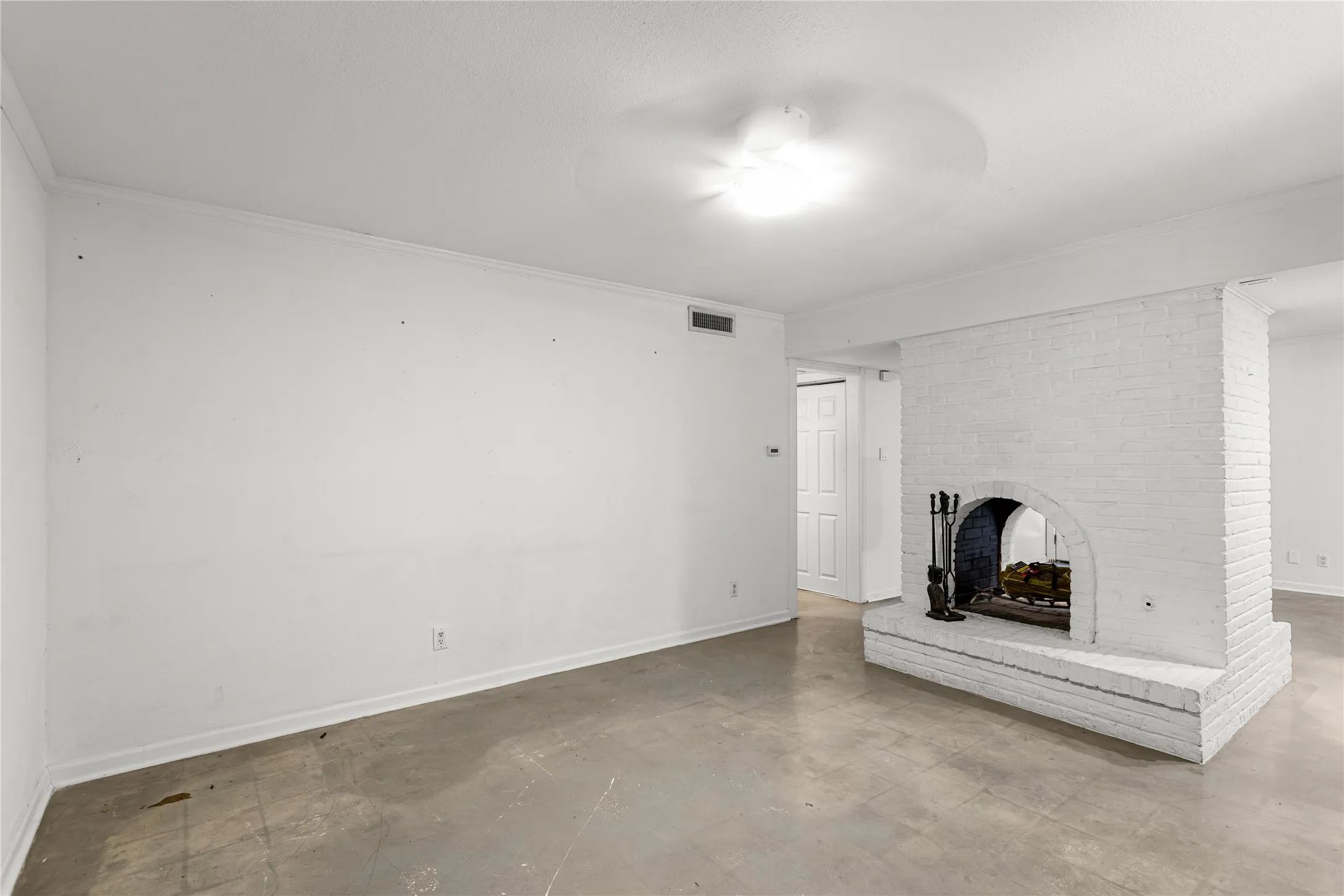 Spacious room featuring a white-painted brick fireplace with a hearth, light-colored flooring, white walls, white baseboards, and a ceiling-mounted light fixture with a fan