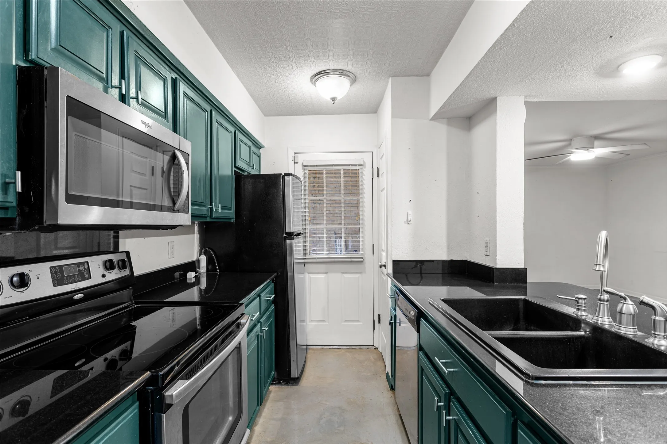 Galley kitchen featuring teal cabinetry, black countertops, stainless steel appliances, and a double basin sink with a chrome faucet