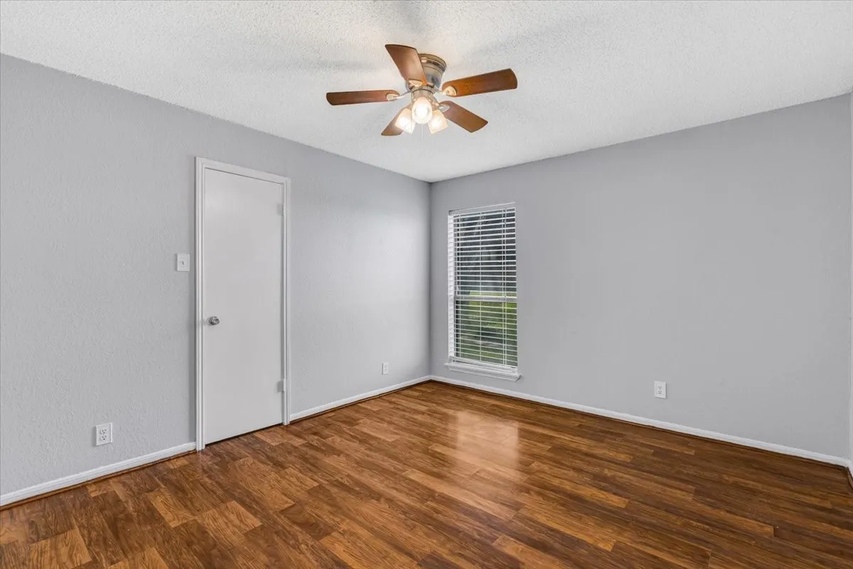 Wood-finish flooring, light gray wall finish, window with blinds, overhead ceiling fan with light, and white interior door