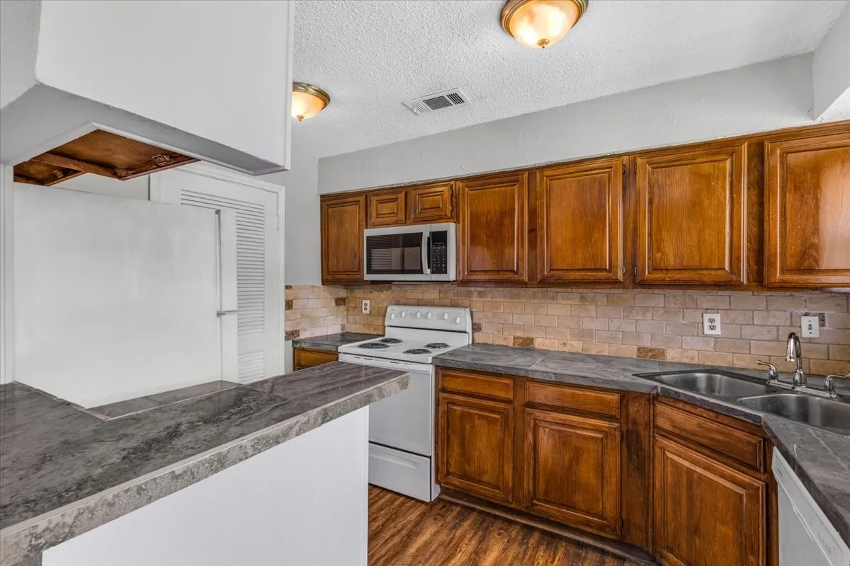 Kitchen featuring rich wood cabinetry, stone-look countertops, a tiled backsplash, wood-finish flooring, and a double basin sink.