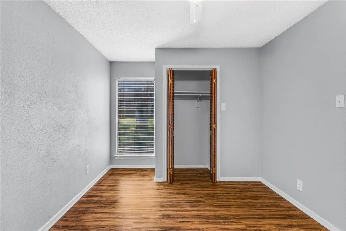 Bright bedroom showcasing warm wood-finish flooring, neutral gray walls, and abundant natural light from a large window with plantation shutters