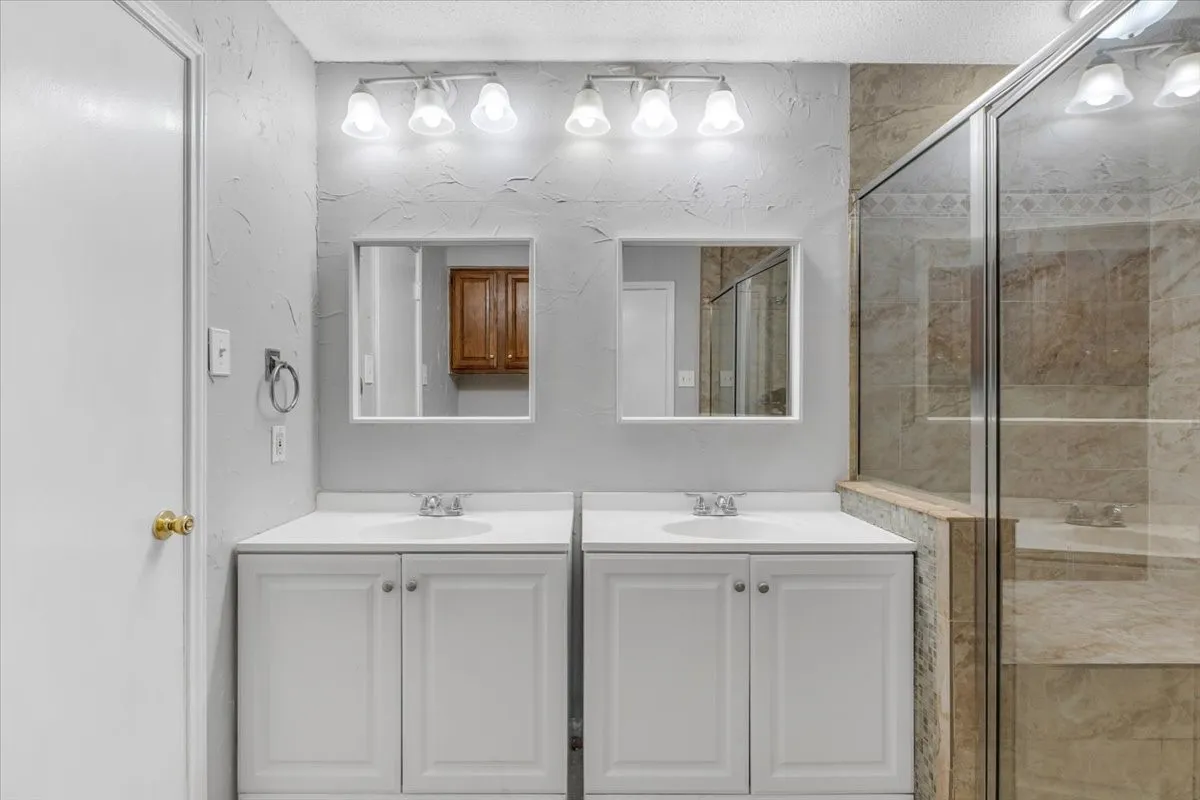Dual white vanities with integrated sinks, individual rectangular mirrors, and a glass-enclosed shower with stone-look tile