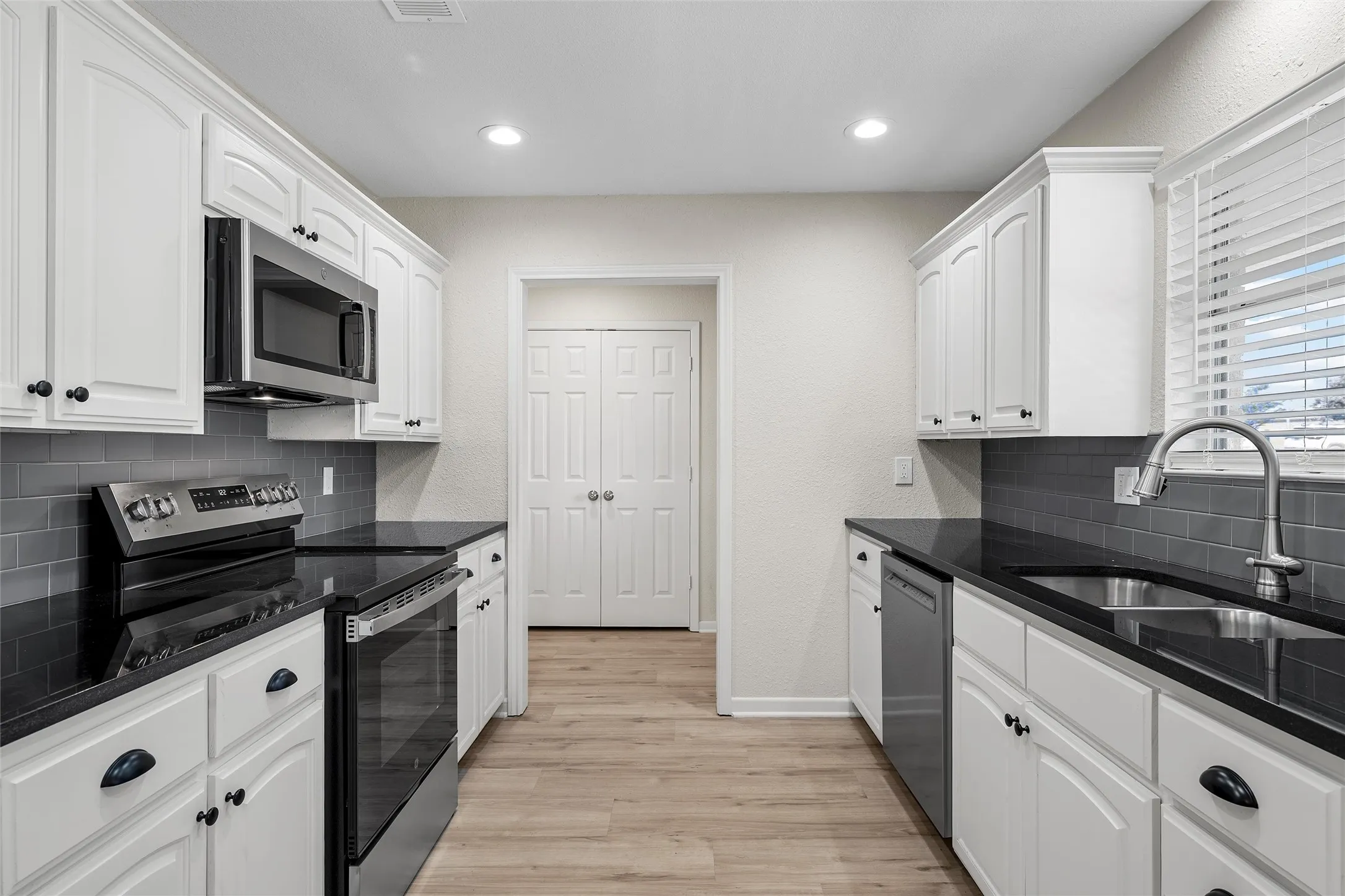 Galley kitchen featuring white cabinetry with dark hardware, dark countertops, a subway tile backsplash, stainless steel appliances, and wood-finish flooring