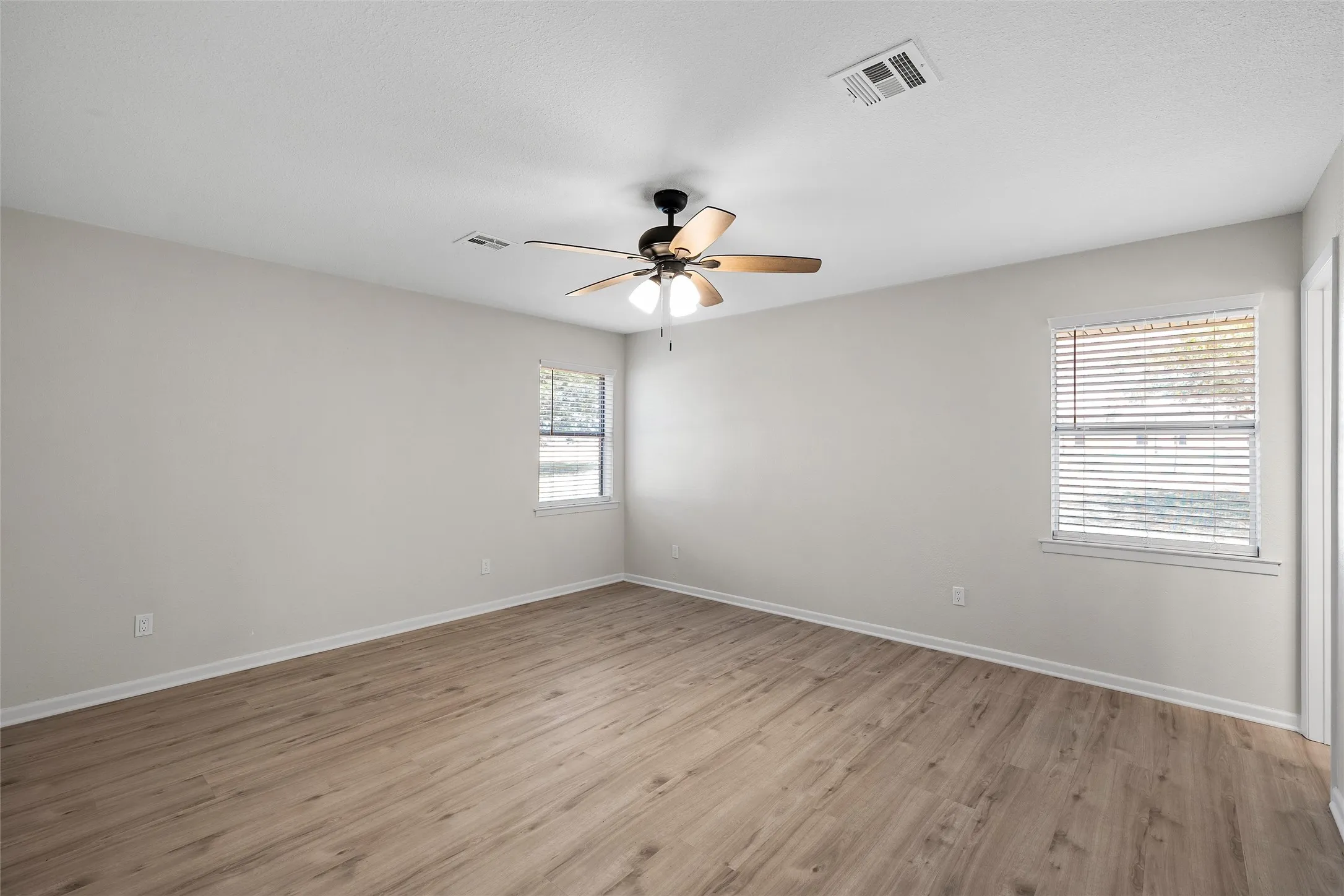 Spacious room featuring wood-finish flooring, two windows with blinds, a ceiling fan with integrated lighting, and neutral wall paint