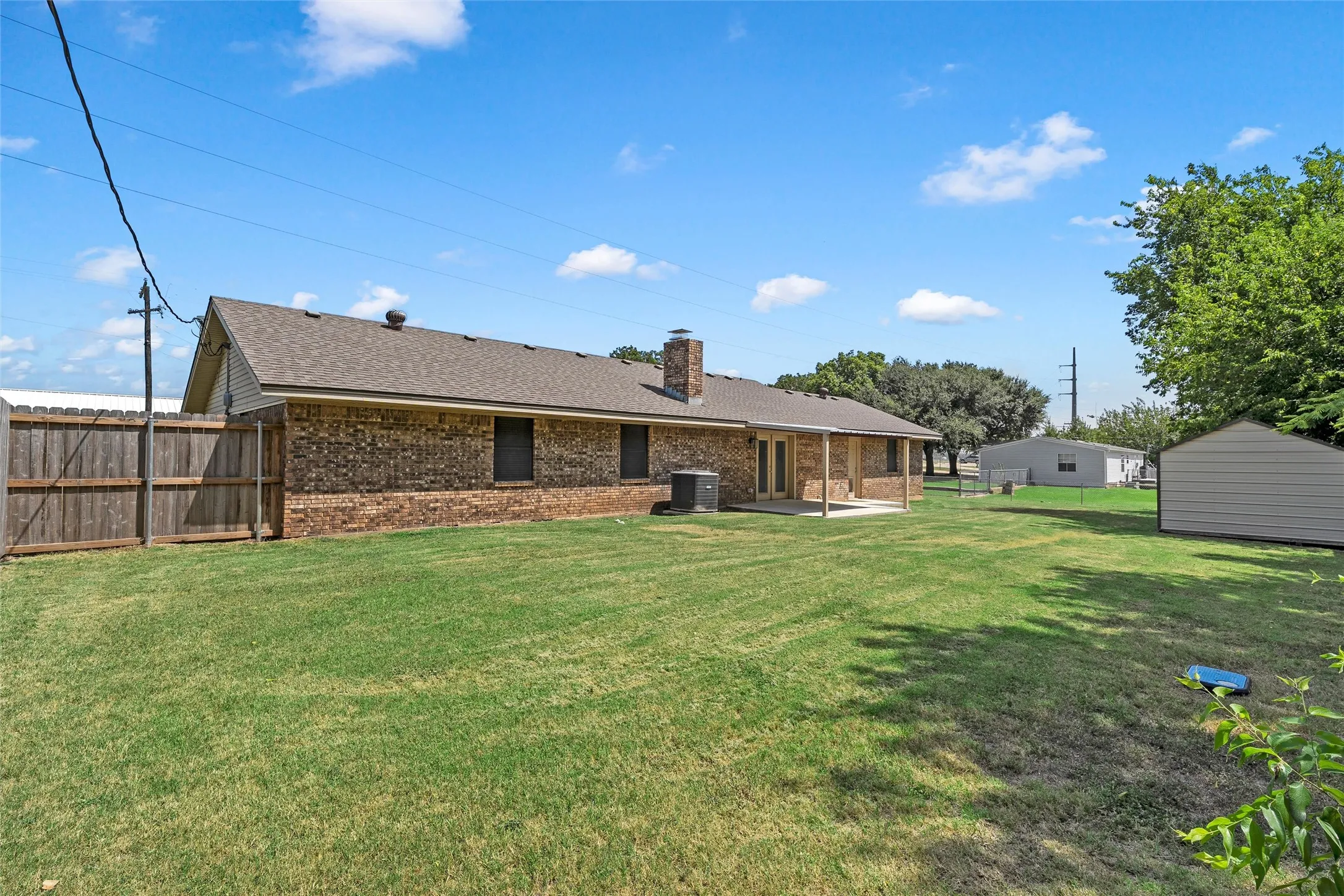 Spacious backyard featuring a large grass lawn, brick exterior, covered patio, and a separate storage shed