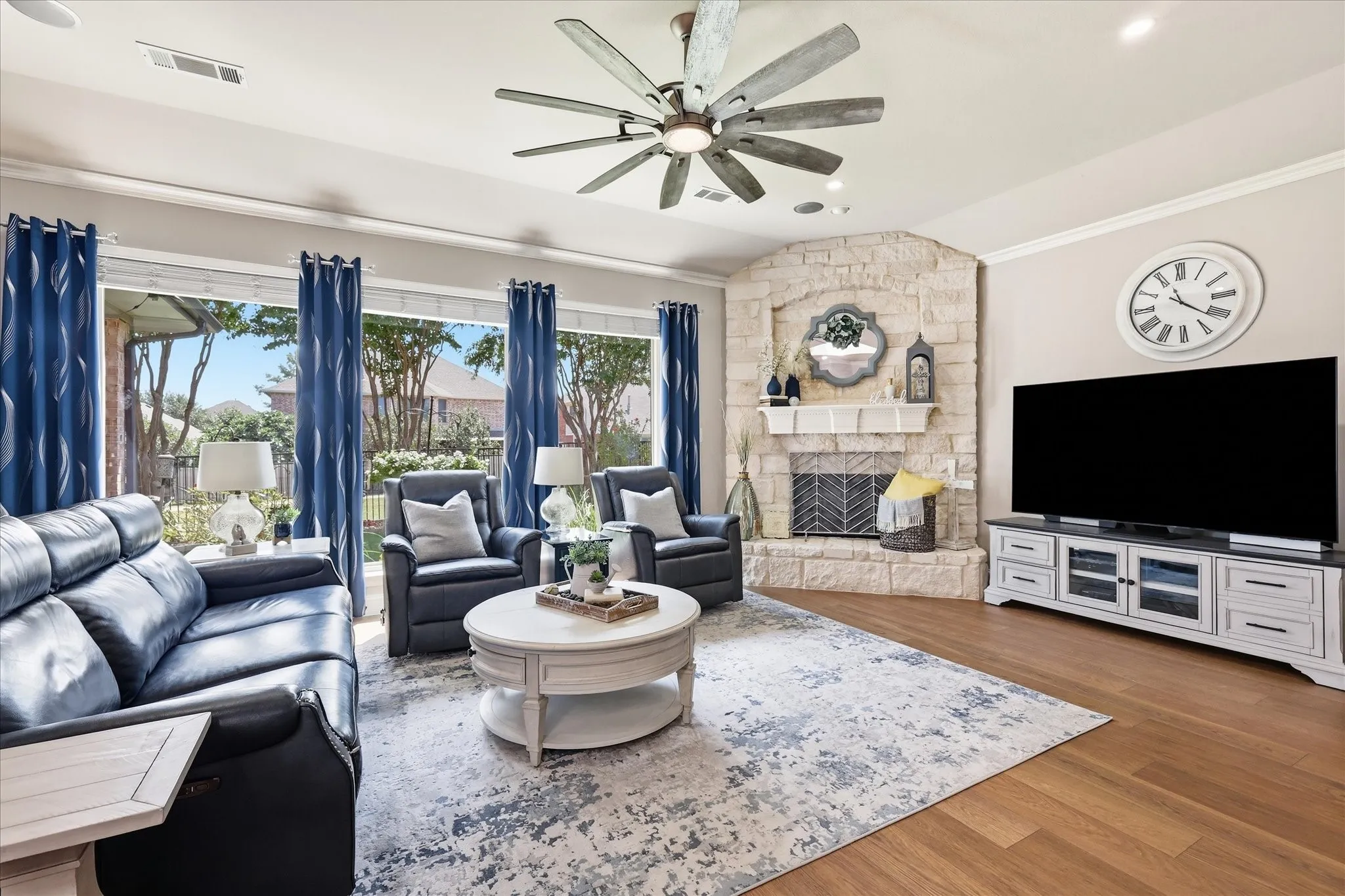 Stone-clad corner fireplace with mantel, ceiling fan with integrated lighting, wood-finish flooring, and expansive windows