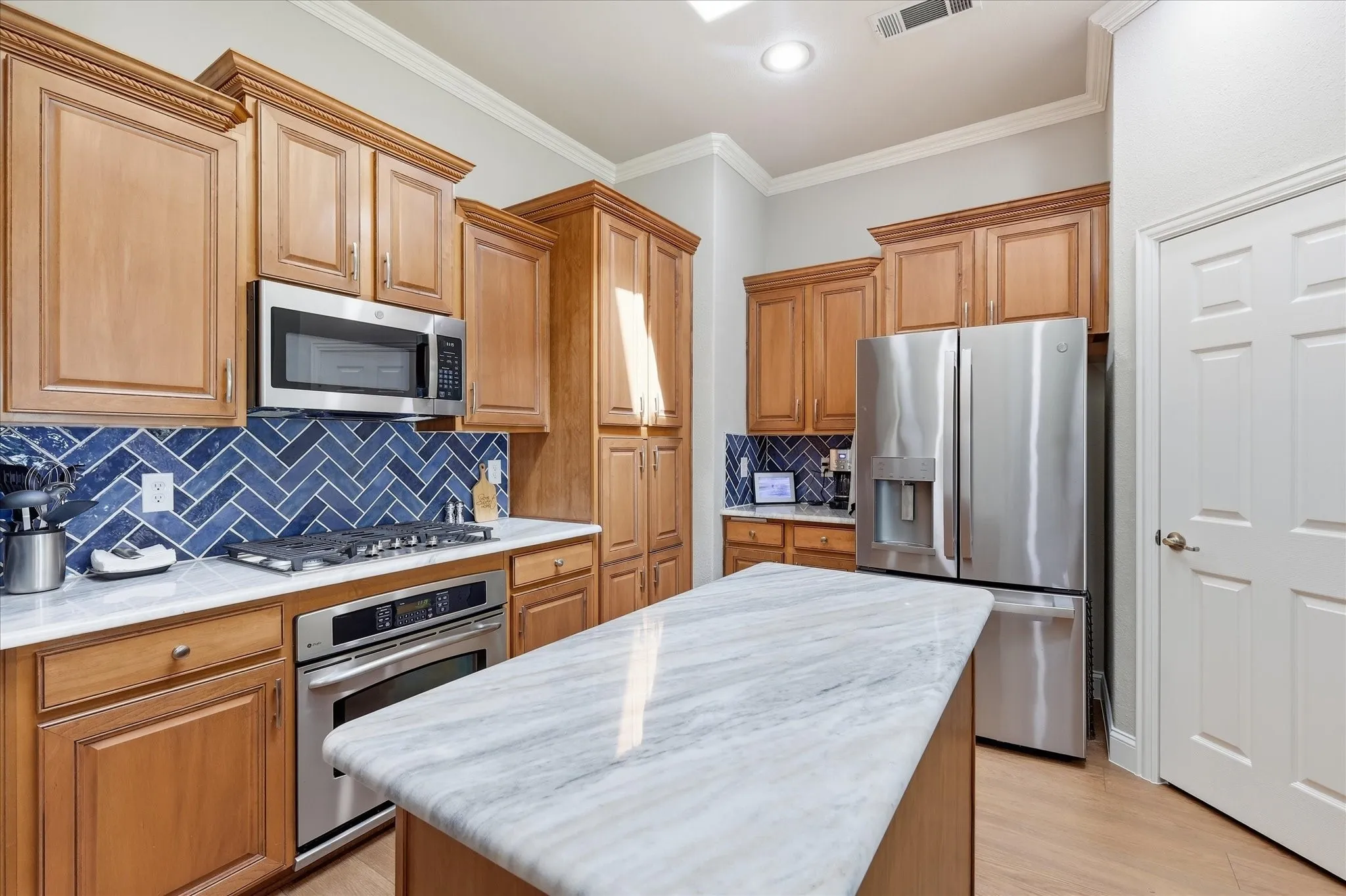 Kitchen featuring wood cabinetry, stainless steel appliances, white countertops, a blue herringbone tile backsplash, and a central island with a white marble-look countertop
