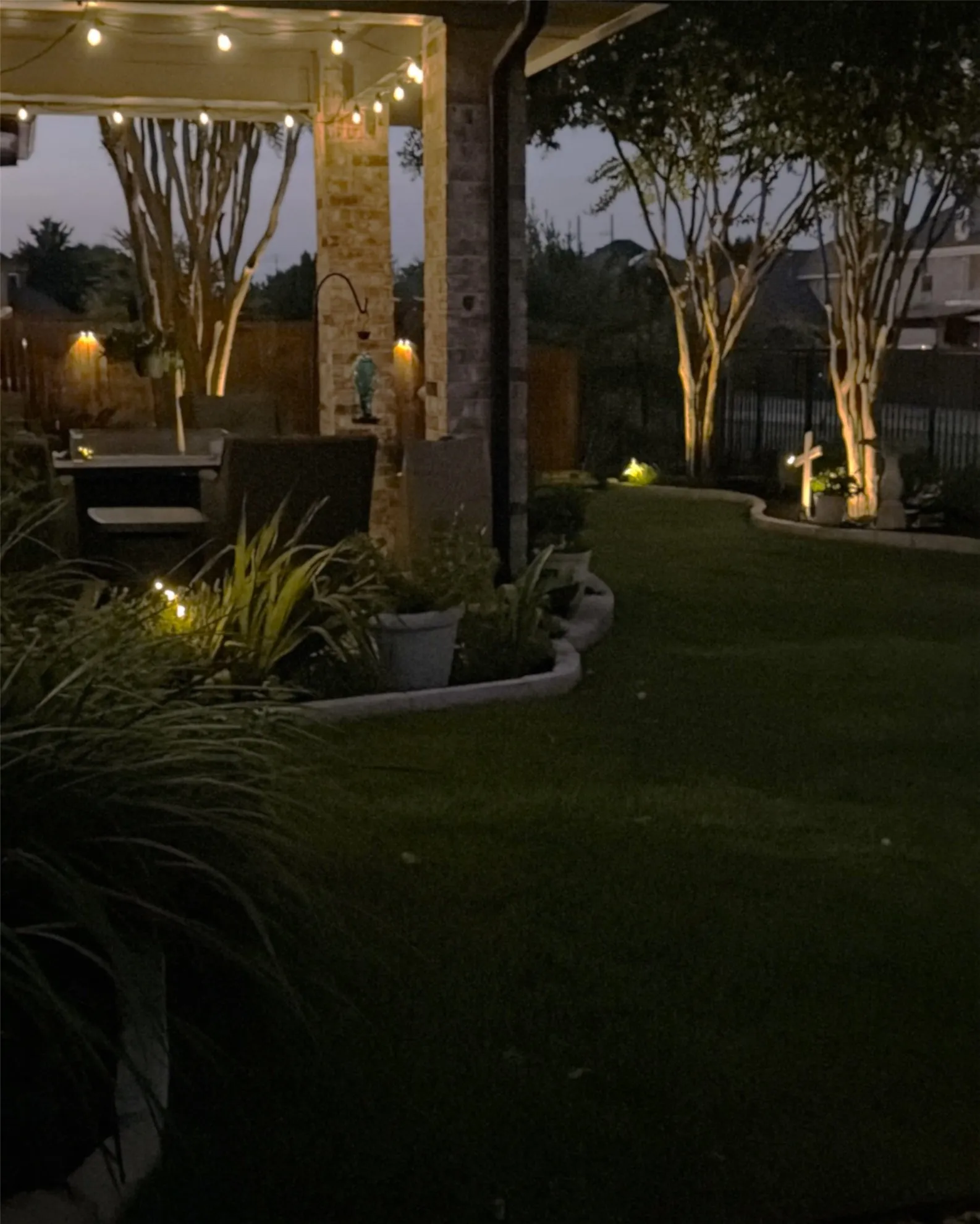 Covered patio with string lighting, stone-clad columns, and a manicured lawn featuring mature trees with accent lighting