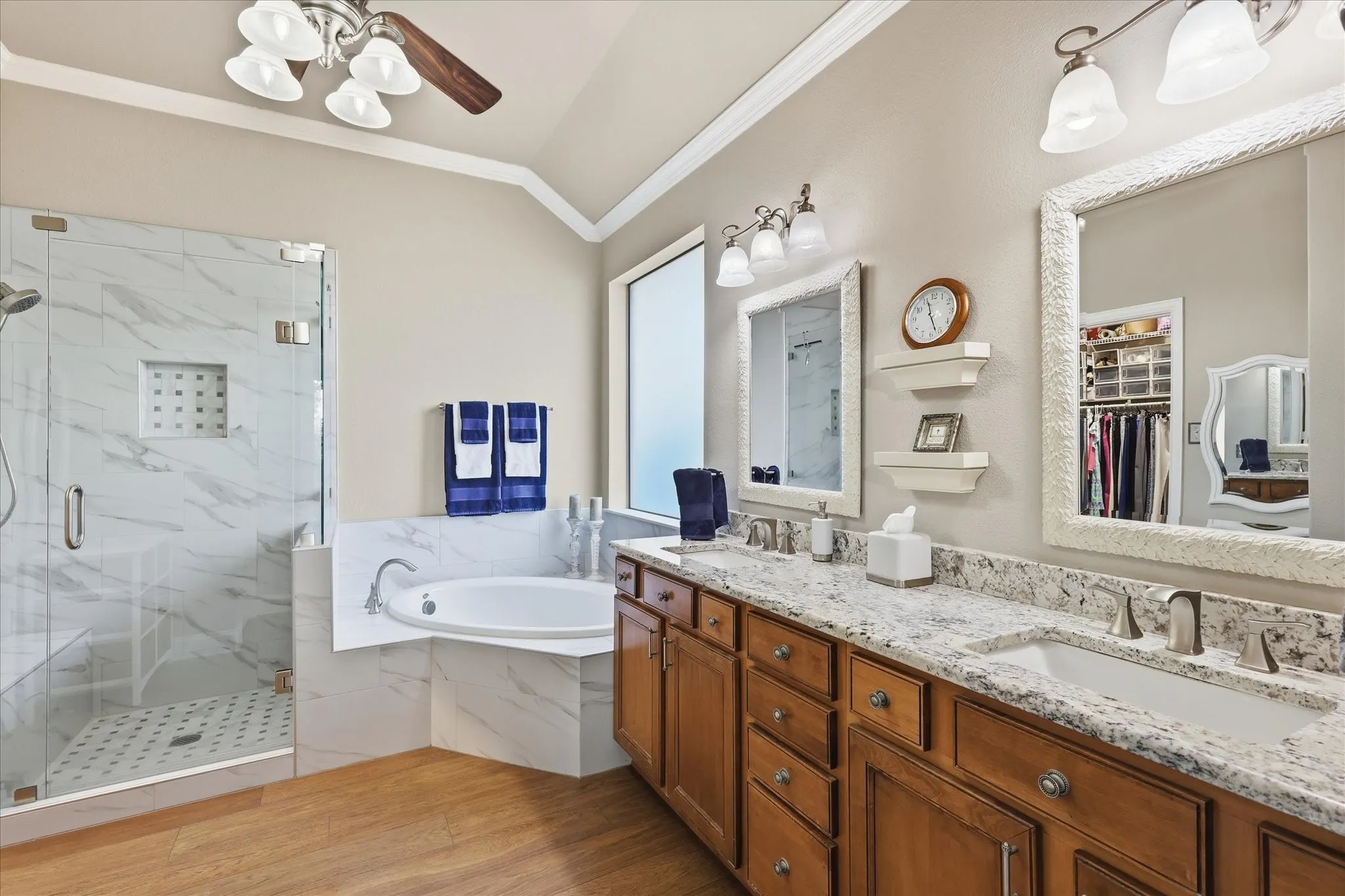 Spacious bathroom featuring a double vanity with granite countertops, wood-finish flooring, a separate walk-in shower with a glass enclosure, and a corner soaking tub