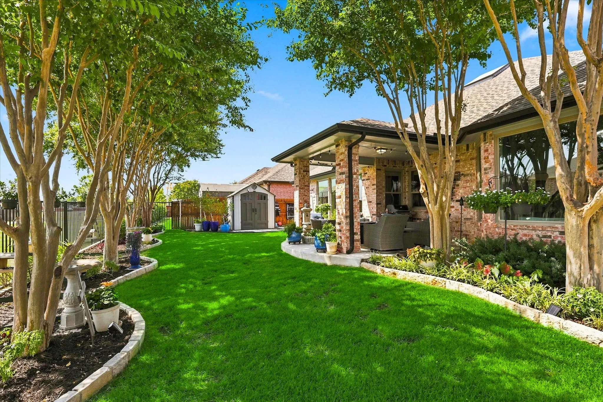 Expansive backyard featuring a manicured lawn, established trees, covered patio, brick exterior, and a detached storage shed