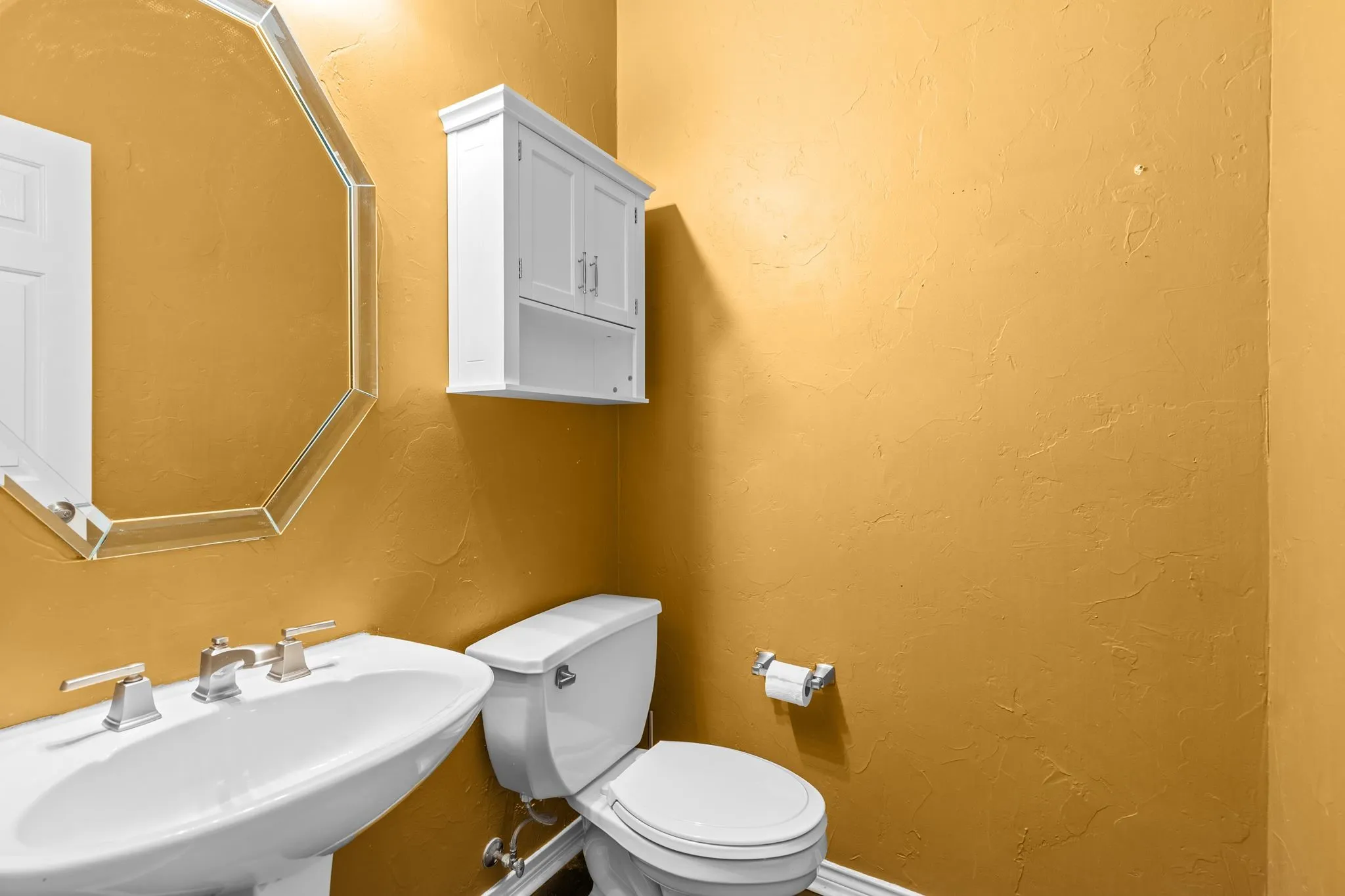 Pedestal sink with brushed nickel faucet, octagonal mirror, and white wall-mounted cabinet