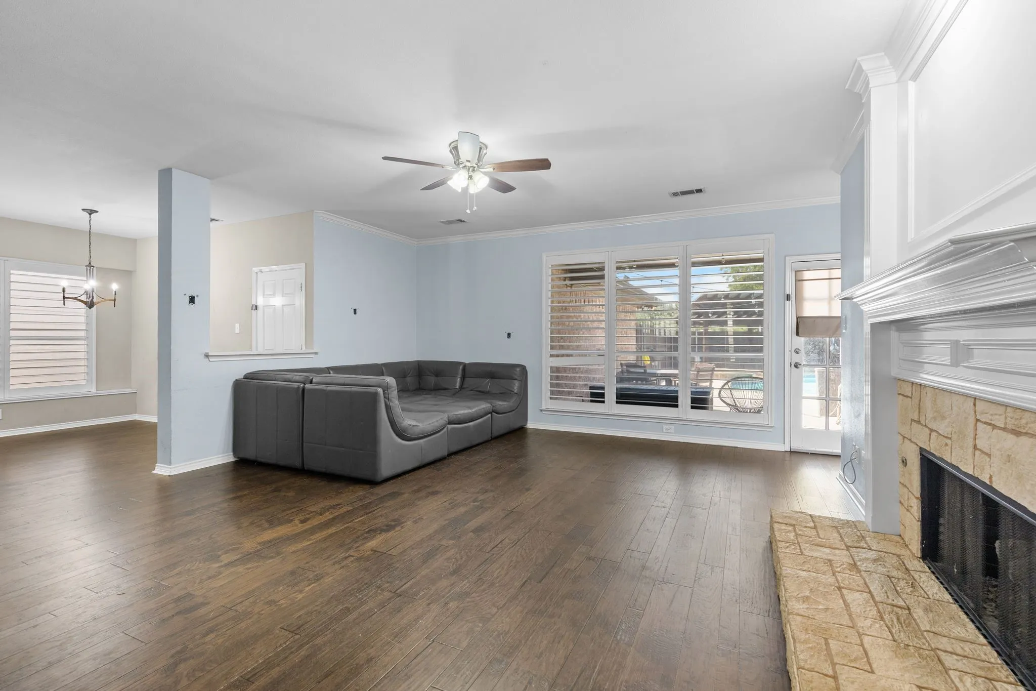 Spacious living area featuring wood-finish flooring, a stone fireplace, large windows with plantation shutters, a ceiling fan, and crown molding