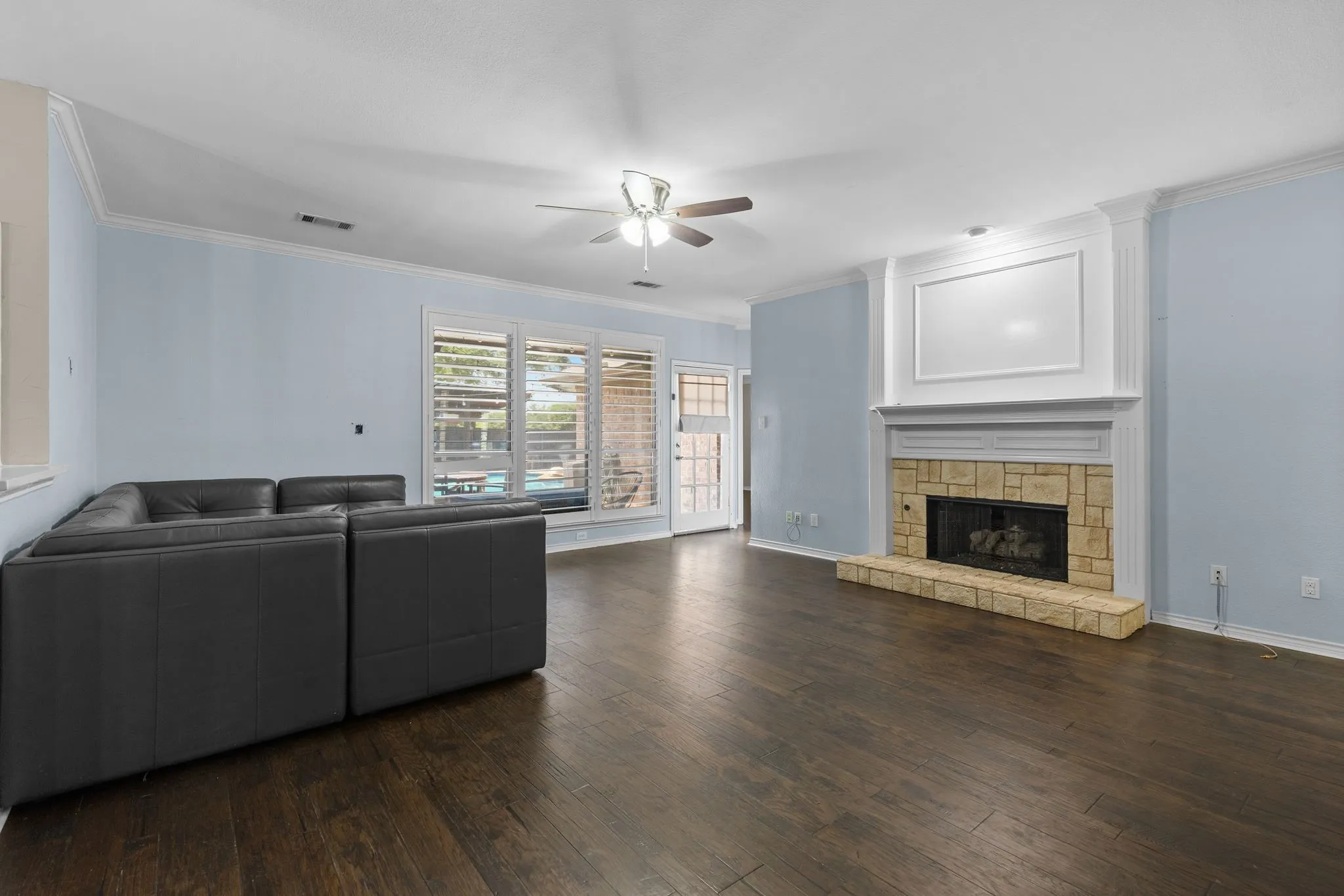 Wood-finish flooring, stone-surround fireplace with mantel, ceiling fan, crown molding, and a transom window with plantation shutters