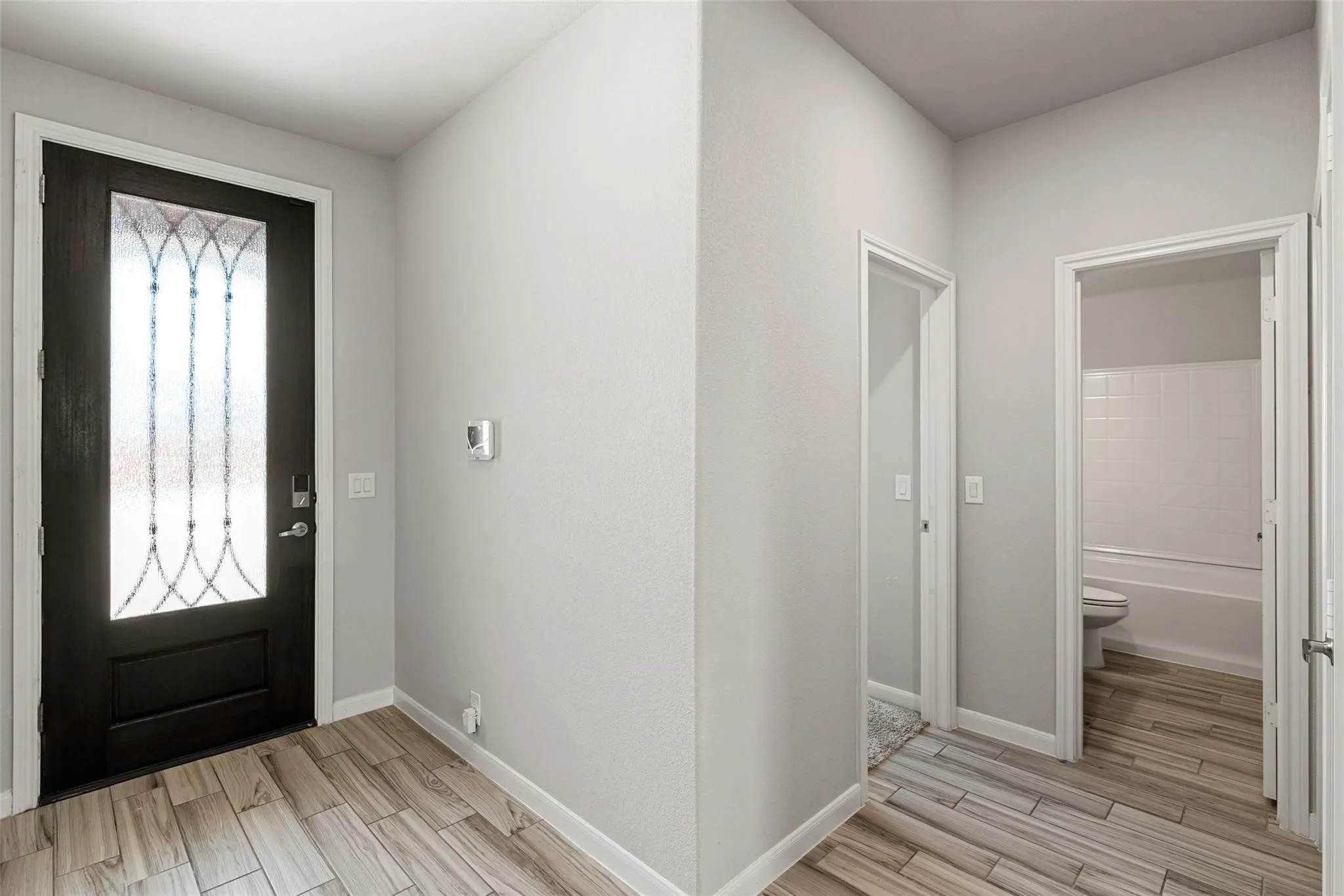 Foyer featuring a dark-finish entry door with decorative glass, wood-finish flooring, light gray walls, white baseboards, and a white interior door leading to a bathroom with a white tiled shower