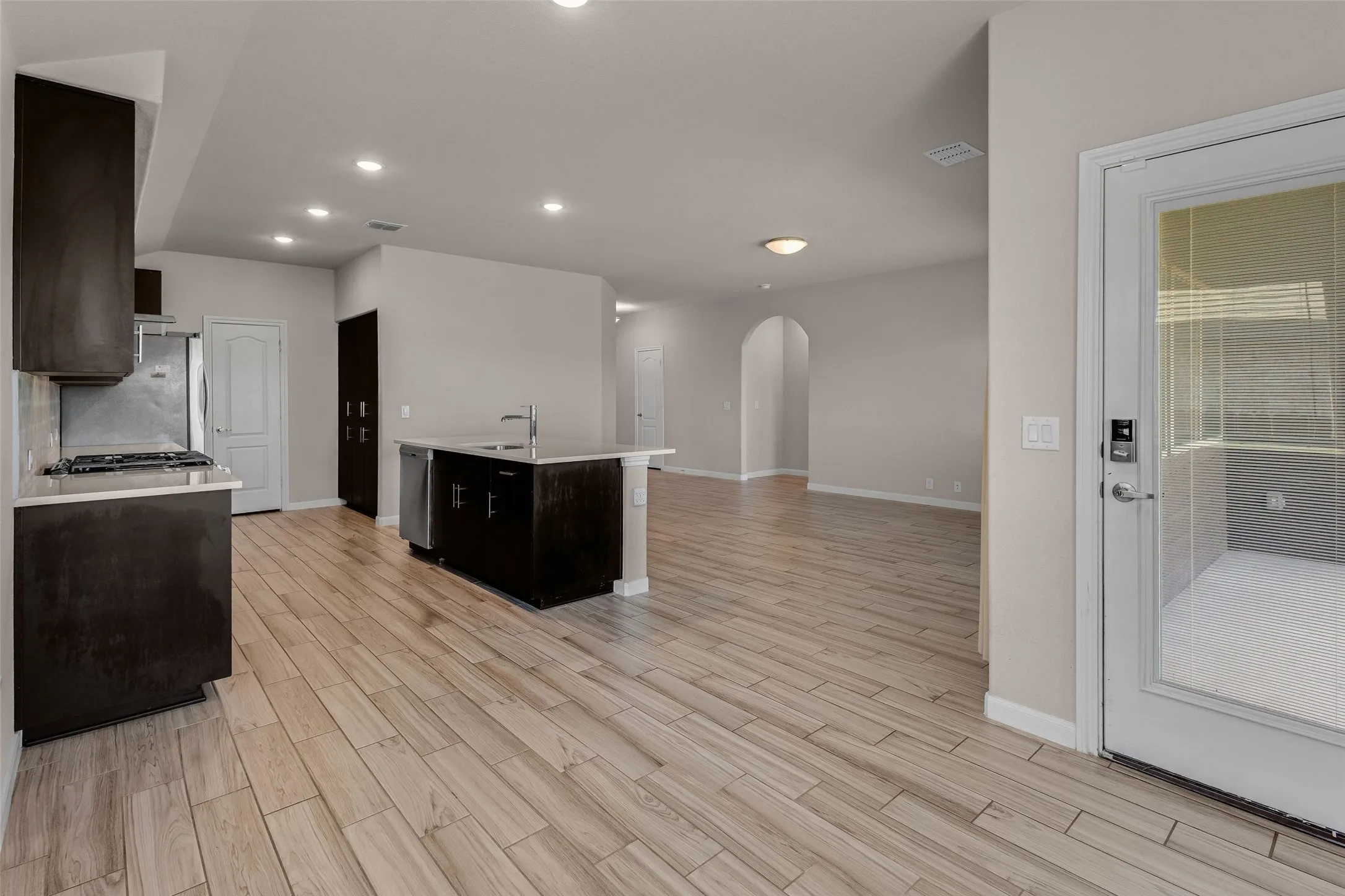Open-concept kitchen featuring dark wood cabinetry, light-toned countertops, a kitchen island with sink, stainless steel appliances, and wood-finish flooring extending into the adjacent living area