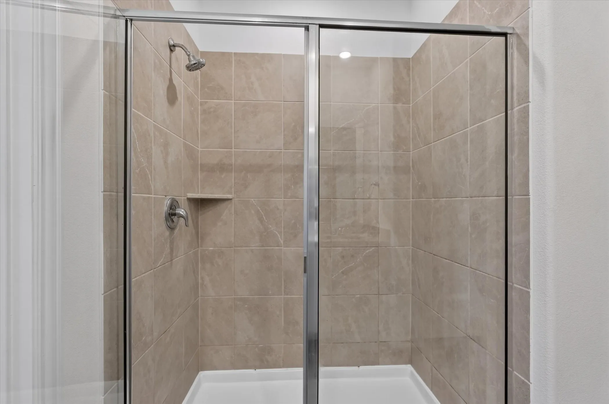 Spa-inspired shower enclosure featuring frameless glass doors with polished chrome hardware, neutral tile surround with linear layout, dual showerheads with individual controls, and a spacious walk-in configuration with built-in shelf niche