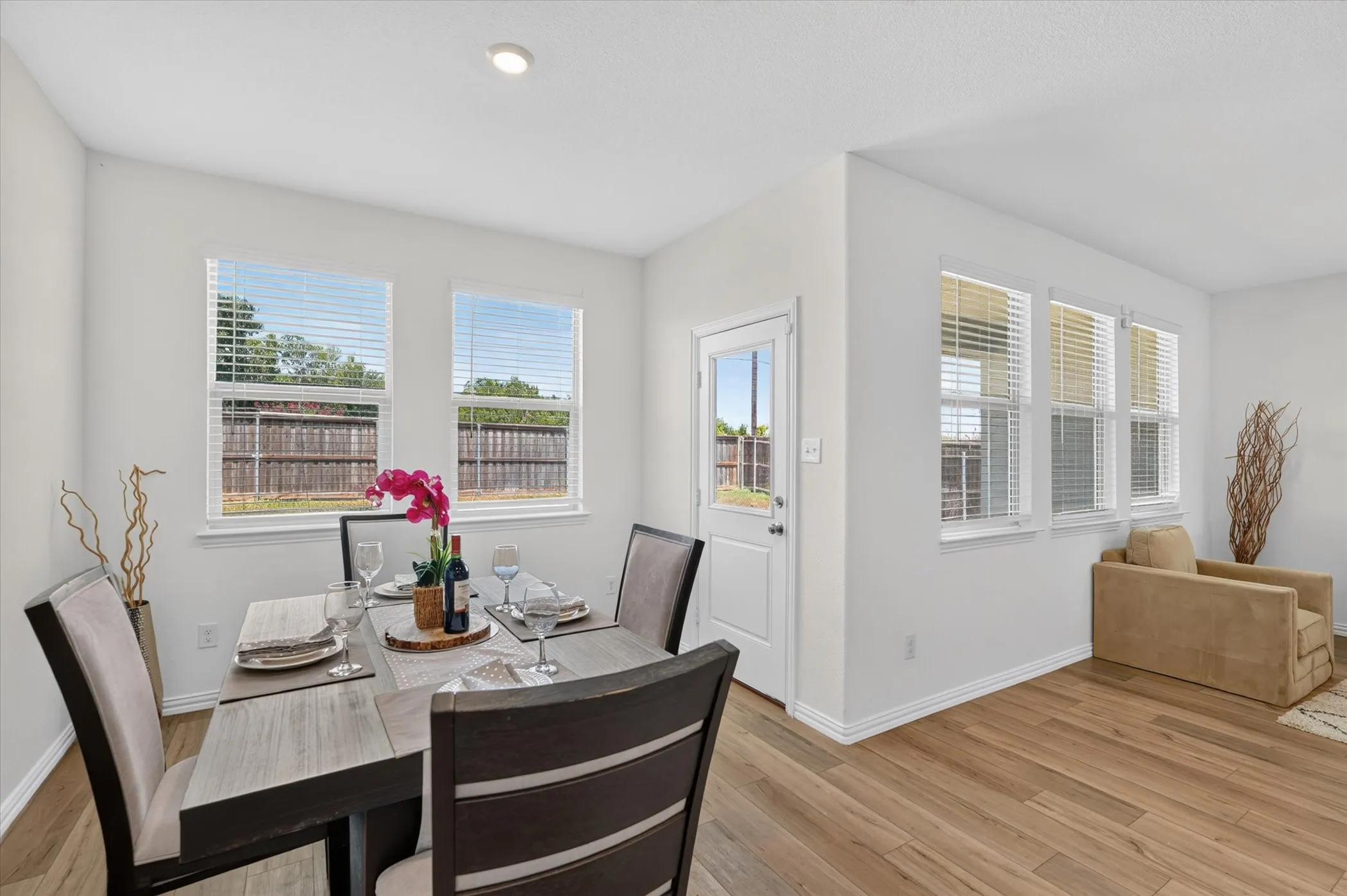 Bright dining area featuring a wood-finish table with upholstered seating, abundant natural light from dual windows, and direct access to an adjacent room through a white entry door